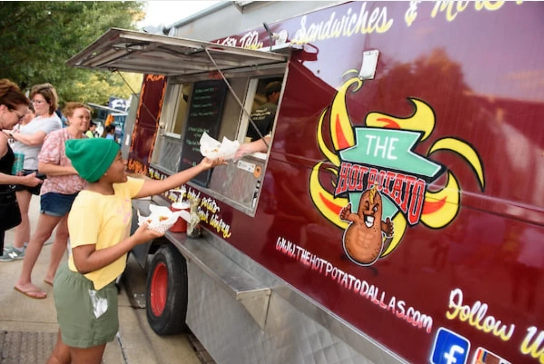 Child in a green beanie and yellow shirt ordering food at a food truck named 'The Hot Potato'.