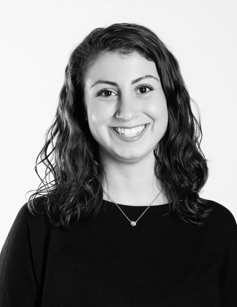 A young woman with curly hair smiling, wearing a black top and a necklace, against a plain background.