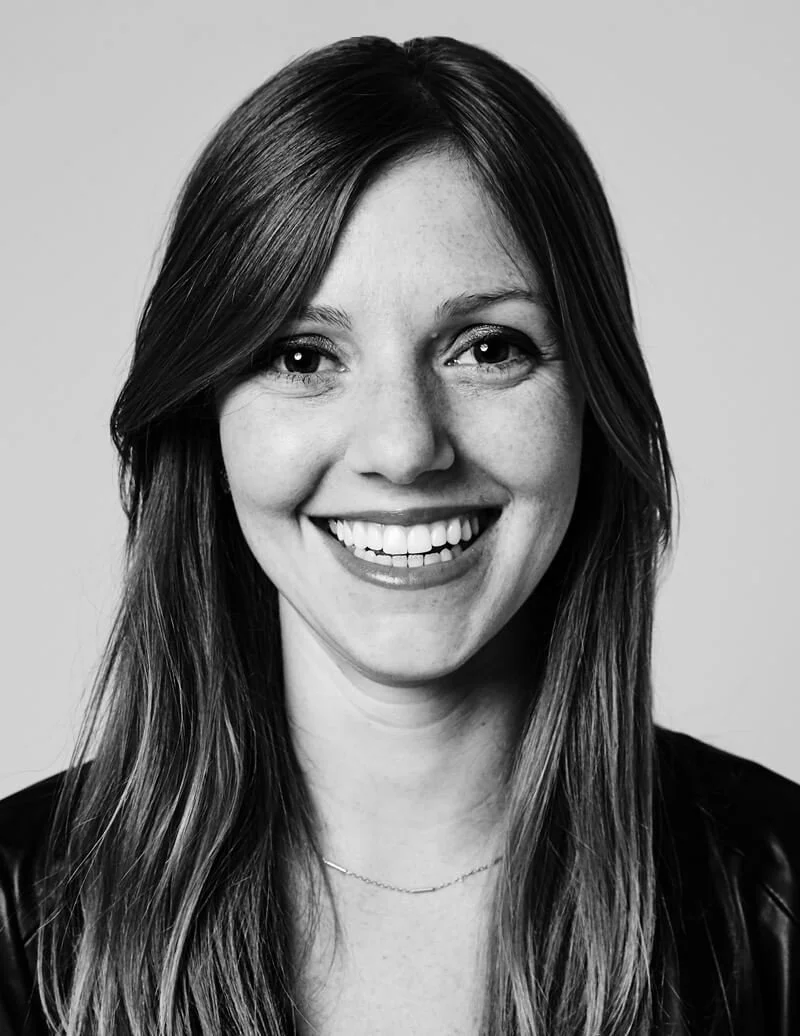 Black-and-white portrait of a smiling woman with long hair, wearing a dark top and a thin necklace.