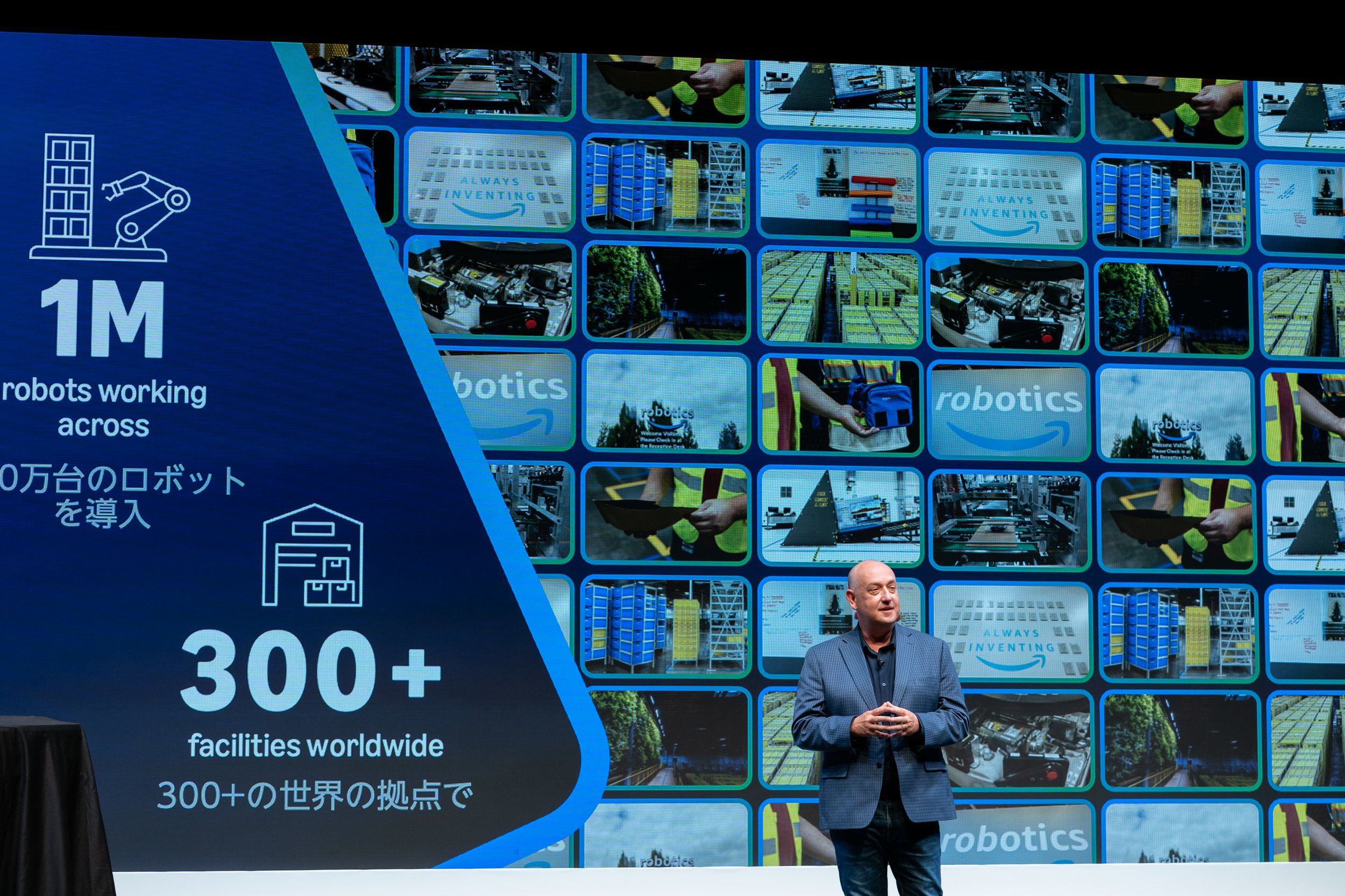 A man in a gray blazer presents on stage with a large screen behind him displaying images of robotics and warehouses. The slide highlights the deployment of over 1 million robots across 300+ worldwide facilities.