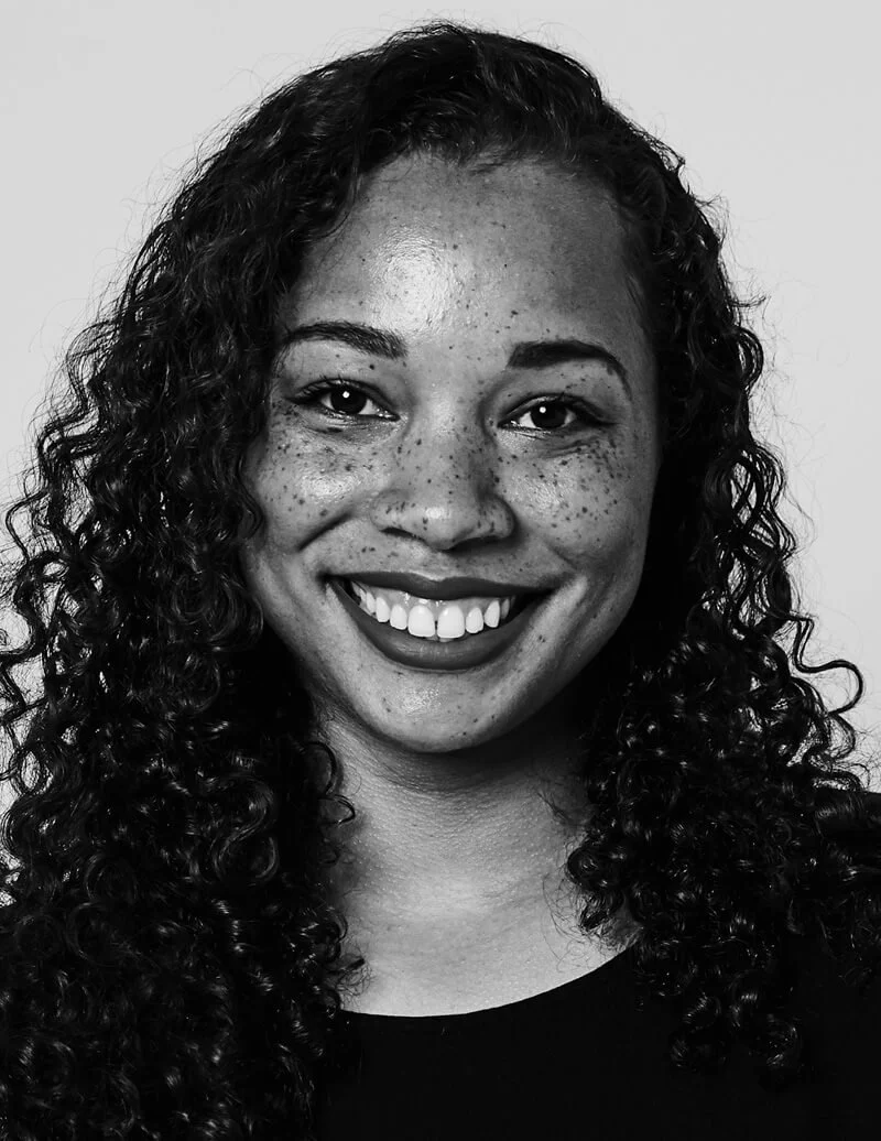 Close-up portrait of a young woman with curly hair, a bright smile showing teeth, and freckles on her face, against a plain background.