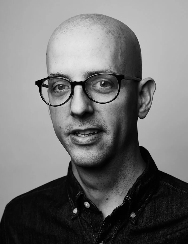 Black and white portrait of a bald man with glasses, wearing a dark button-up shirt, smiling slightly, against a plain background.