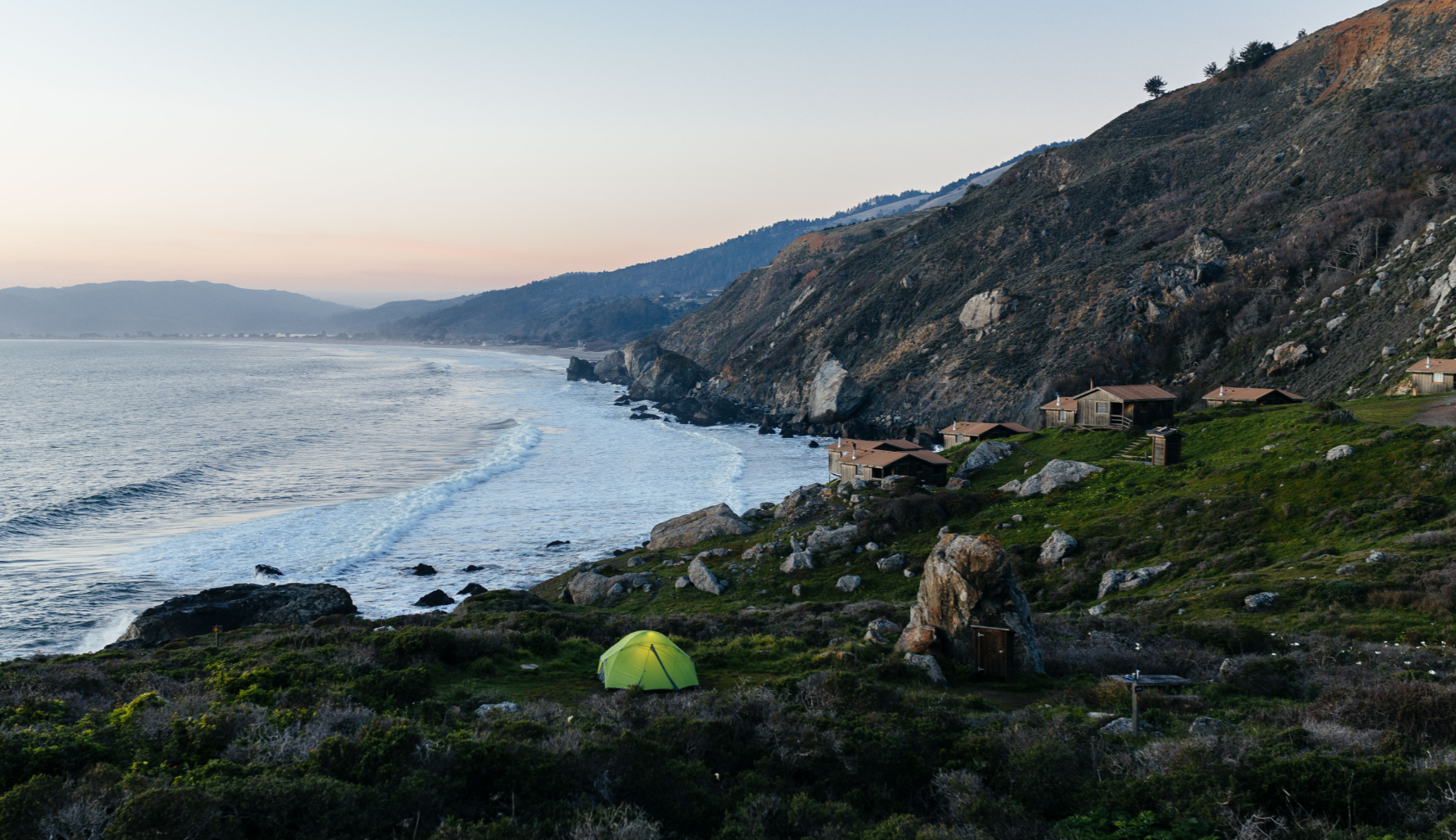 A scenic coastal landscape at dusk with a yellow tent on green grass, rocky cliffs with houses, rolling hills, and ocean waves.
