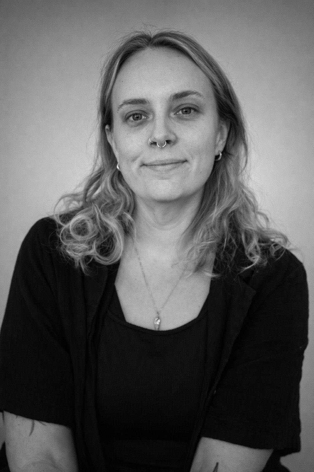 Black and white portrait of a woman with shoulder-length curly blonde hair, wearing large black glasses, a nose ring, and a dark top, smiling at the camera.