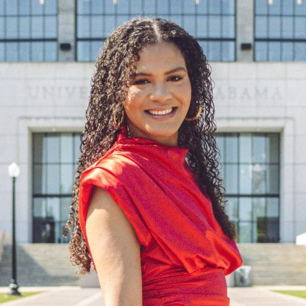 Young woman with curly hair smiling, wearing a red dress and gold hoop earrings, standing outside in front of a government building.