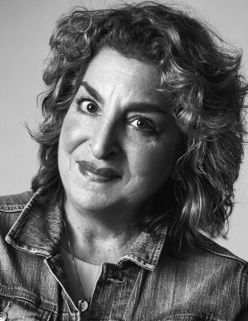 A black-and-white portrait of a woman with curly short hair, wearing a denim jacket and a necklace.
