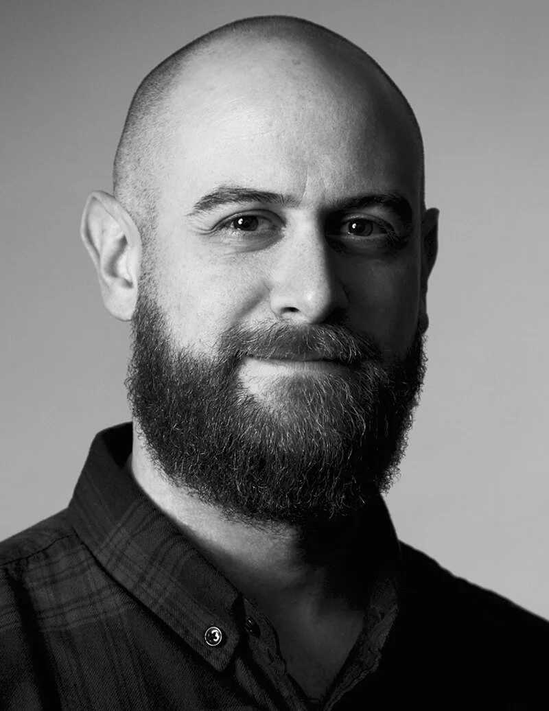 Black and white portrait of a bearded man with a shaved head wearing a dark plaid shirt.