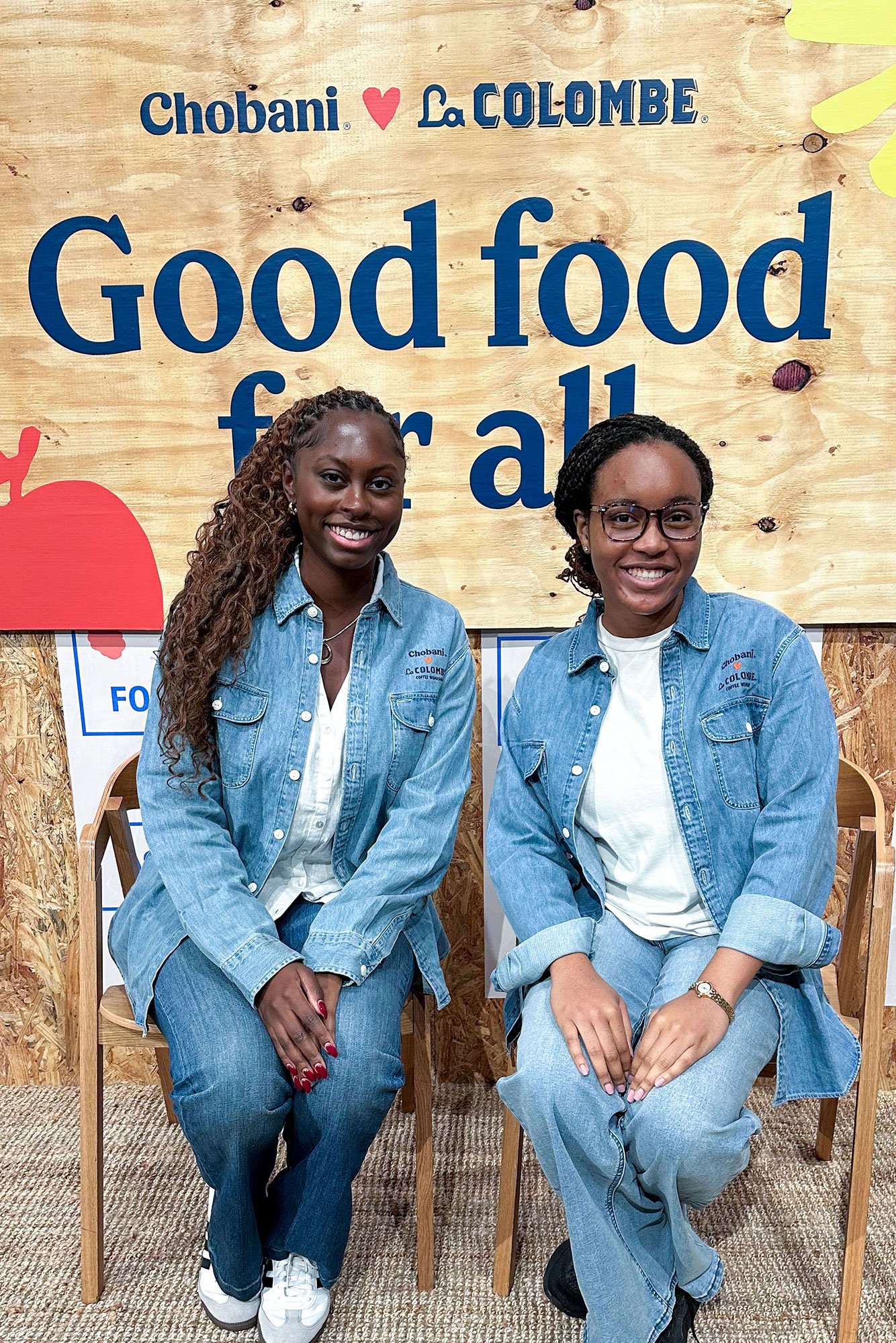Two women sitting on wooden chairs, smiling, wearing denim jackets and jeans, in front of a sign that reads 'Good food for all' with a background of wood paneling.