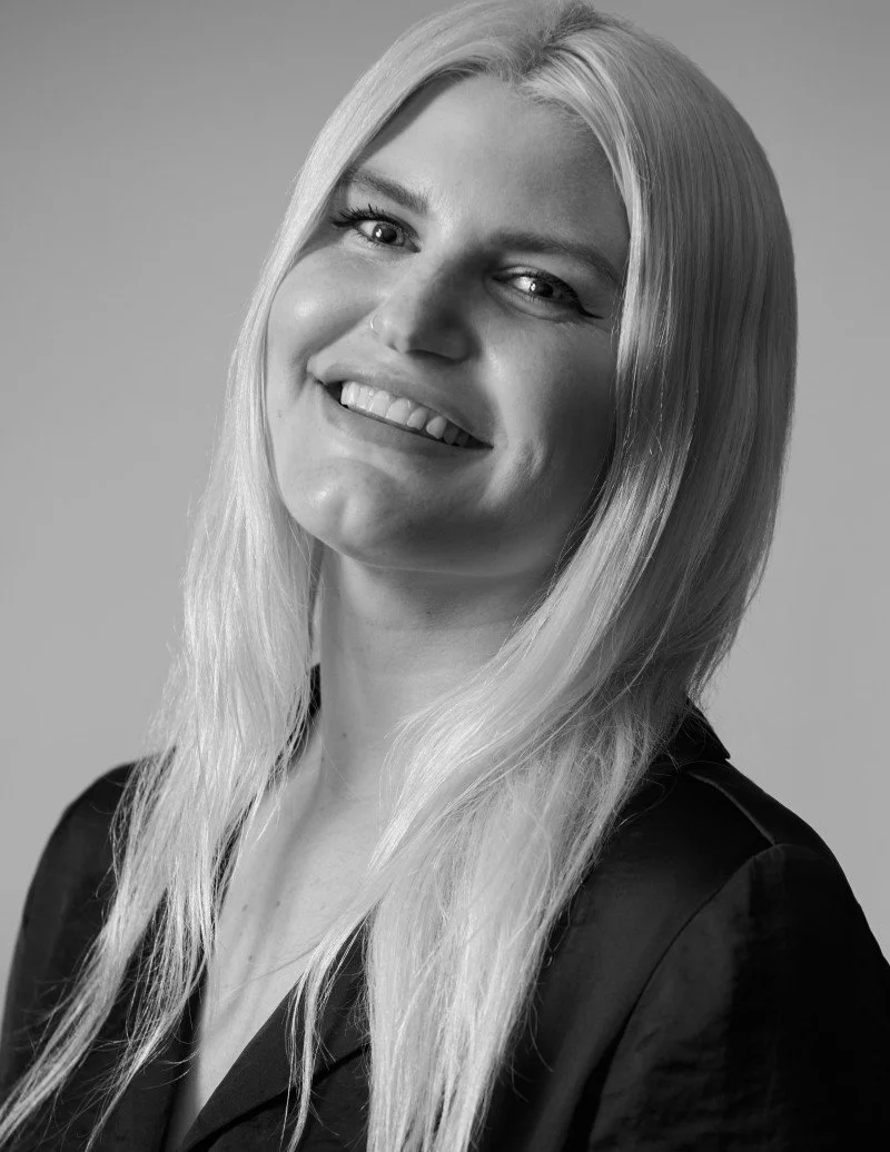 A smiling woman with long blonde hair, wearing a dark top, in a black and white portrait.