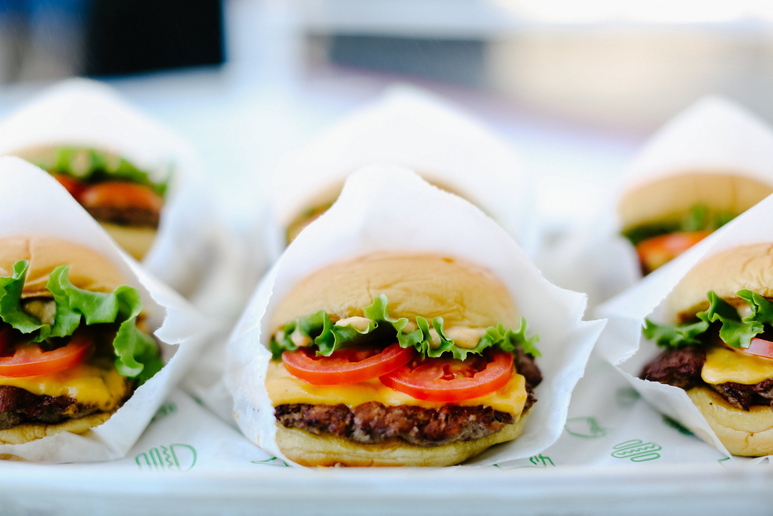 Close-up of several cheeseburgers wrapped in white paper, with lettuce, tomato, cheese, and beef patties visible.