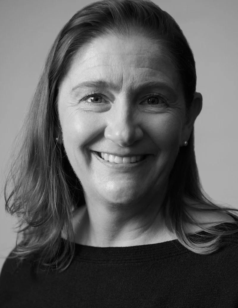A black and white portrait of a smiling woman with shoulder-length hair, wearing a dark top and small earrings.