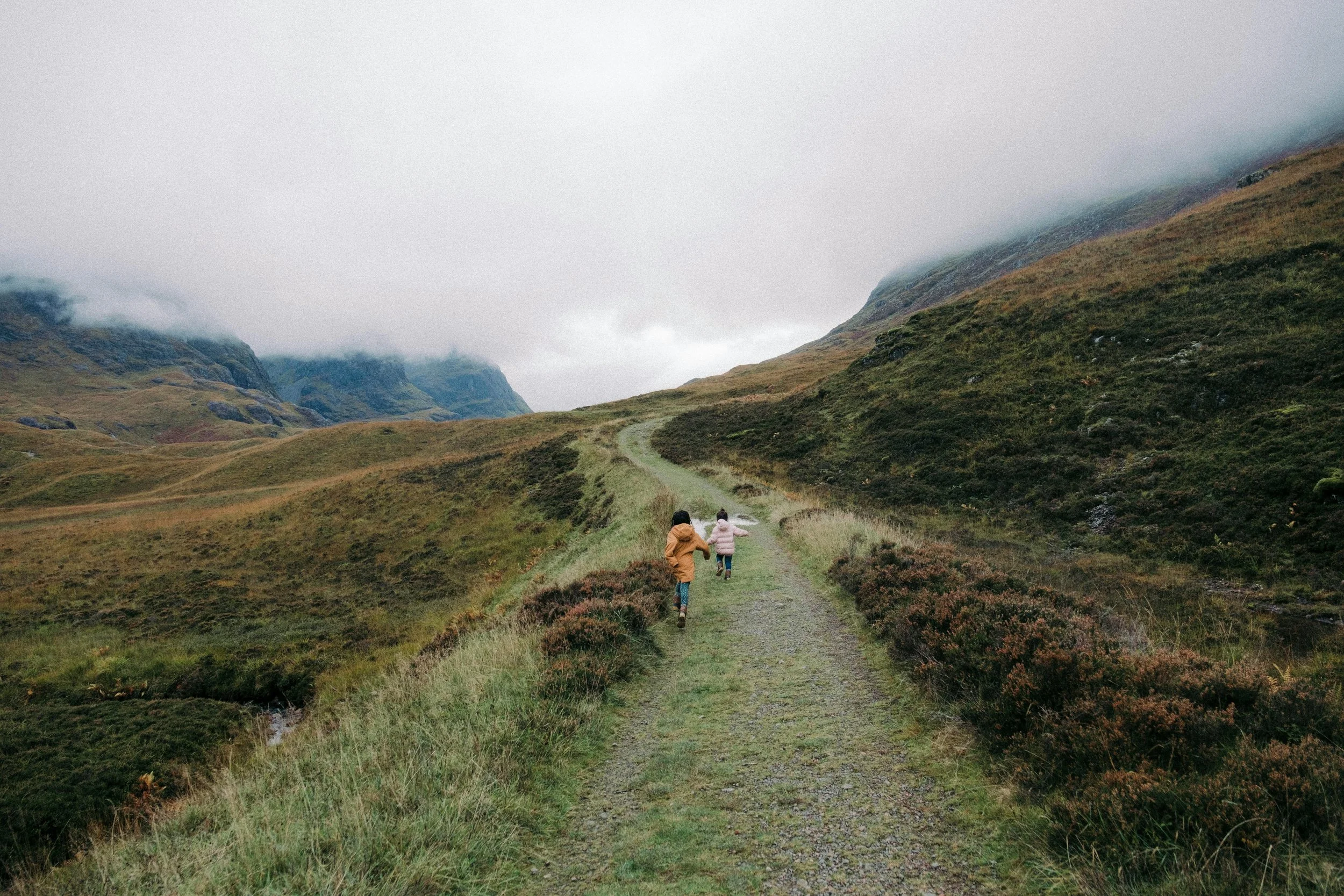 Two children running on a trail through green hilly landscape with mountains and fog in the background.