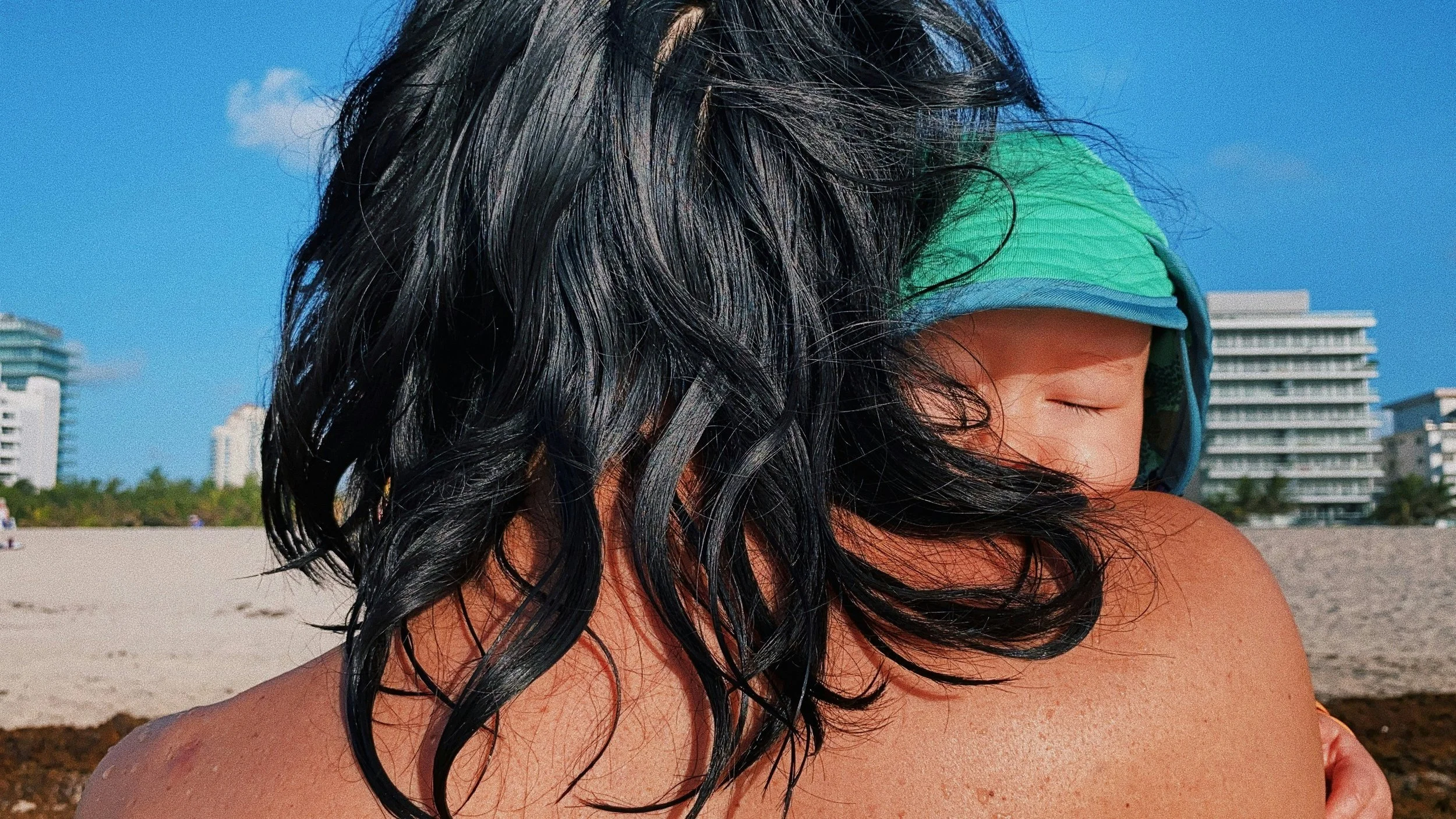 A person with long, black, wet hair cuddling a child wearing a green and blue cap at the beach with buildings in the background.