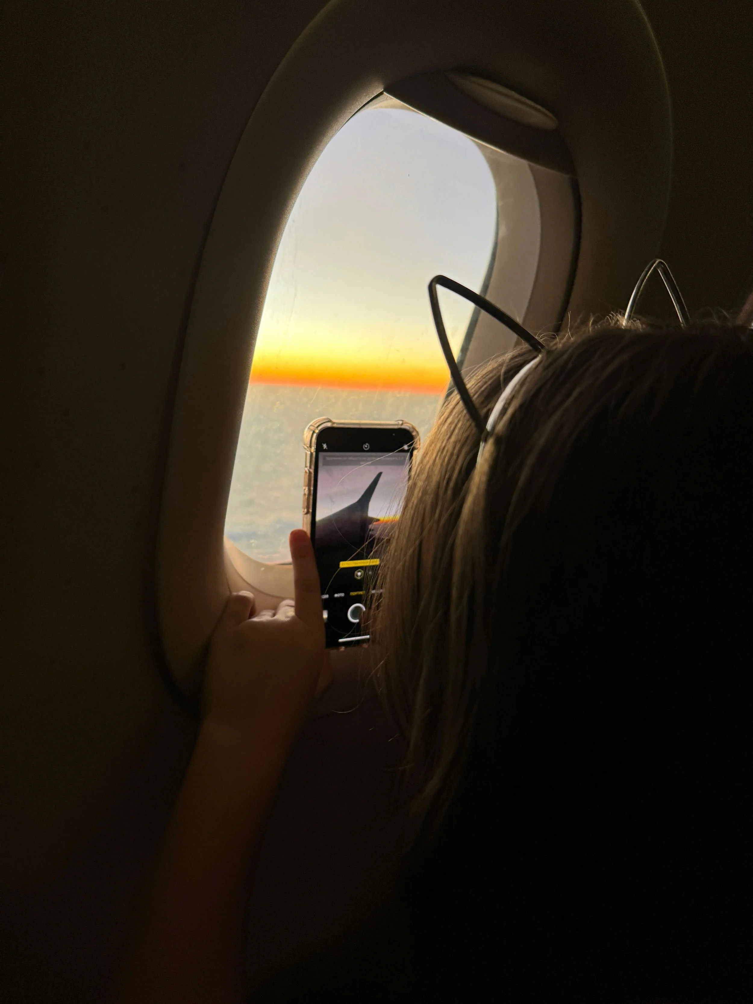 Photo taken from inside an airplane showing a passenger with glasses taking a selfie of the airplane window at sunset, with a view of the sky and horizon outside.