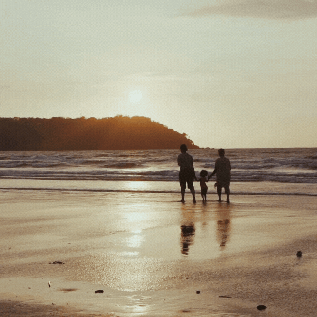 Three people, two adults and a child, holding hands and standing on the beach at sunset, with the ocean and a distant hill in the background.