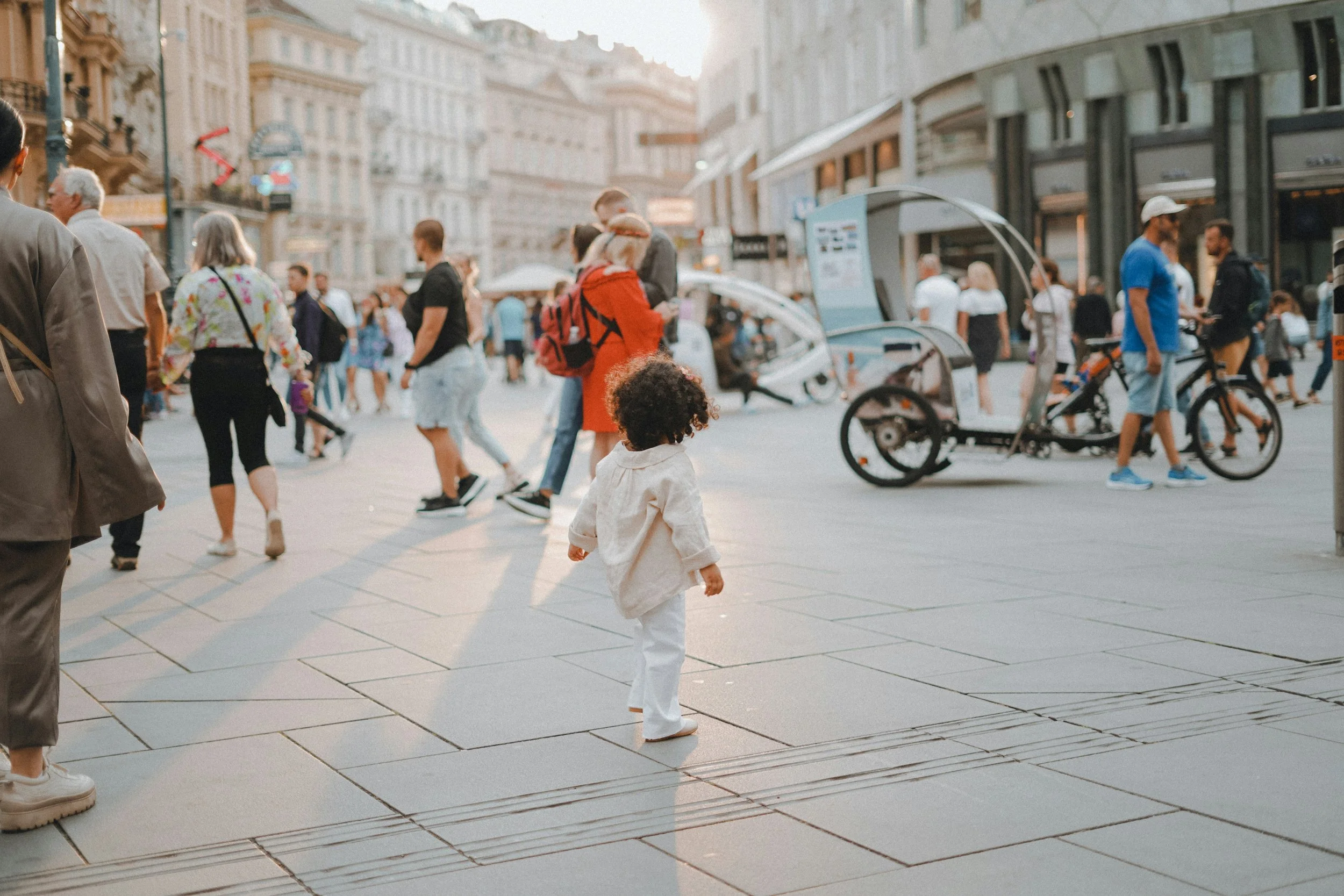 People walking and cycling on a busy city street in the late afternoon or early evening, with buildings and storefronts lining the sidewalks.