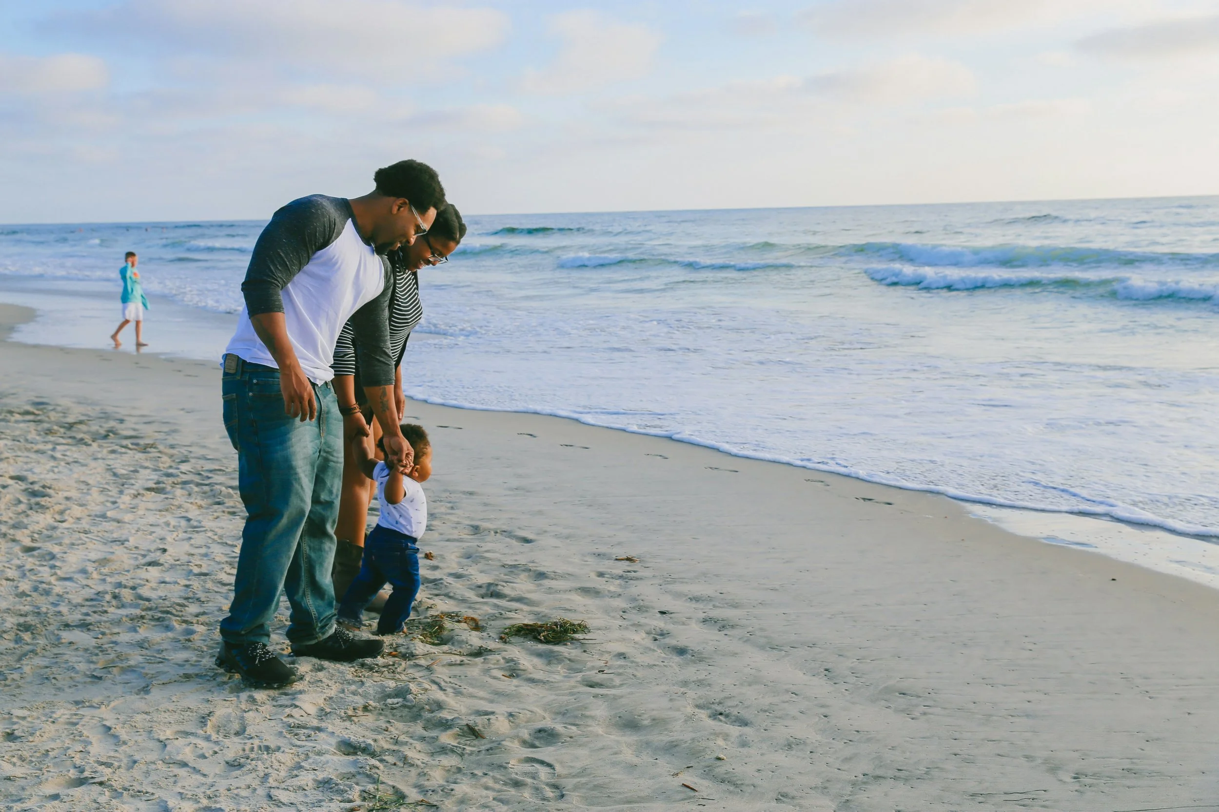 Three people walking along the shoreline at the beach, one man and two children, with one child holding hands with the man. There is another person in the background walking closer to the water, under a partly cloudy sky.