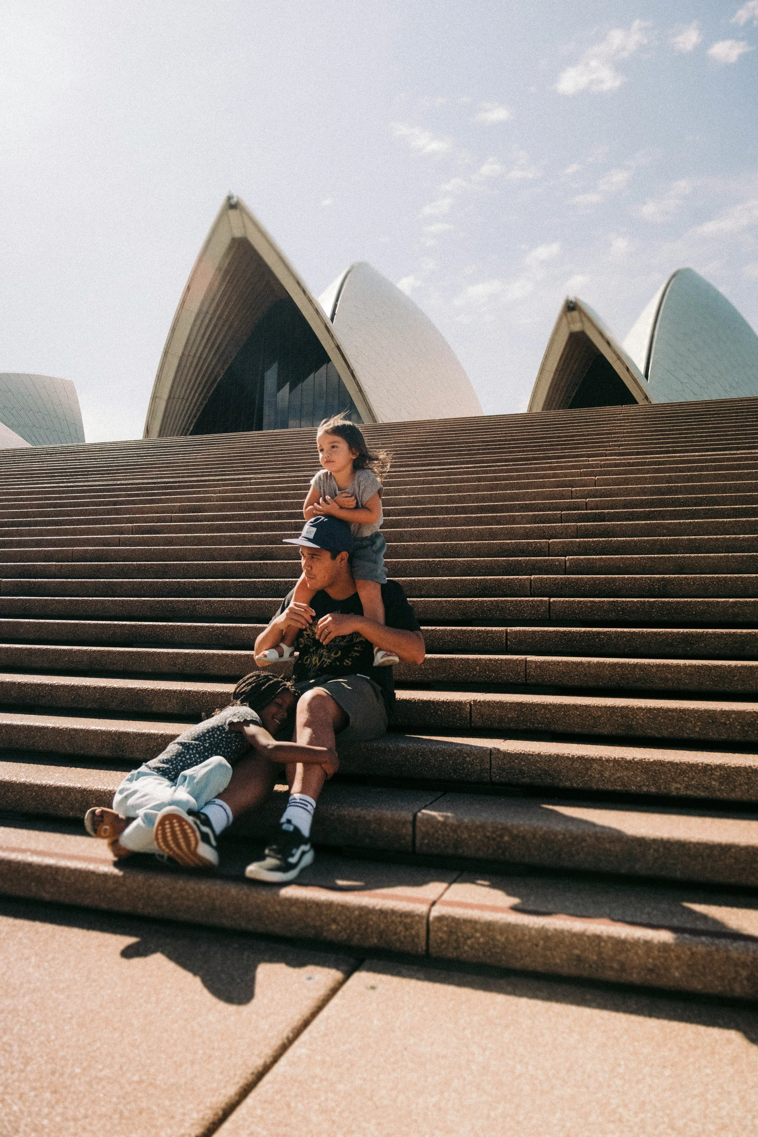 Four children sitting and lying on the steps in front of the Sydney Opera House, Australia, during daylight. The children are sitting in various relaxed poses, with the iconic sail-like structures of the Opera House in the background under a partly cloudy sky.