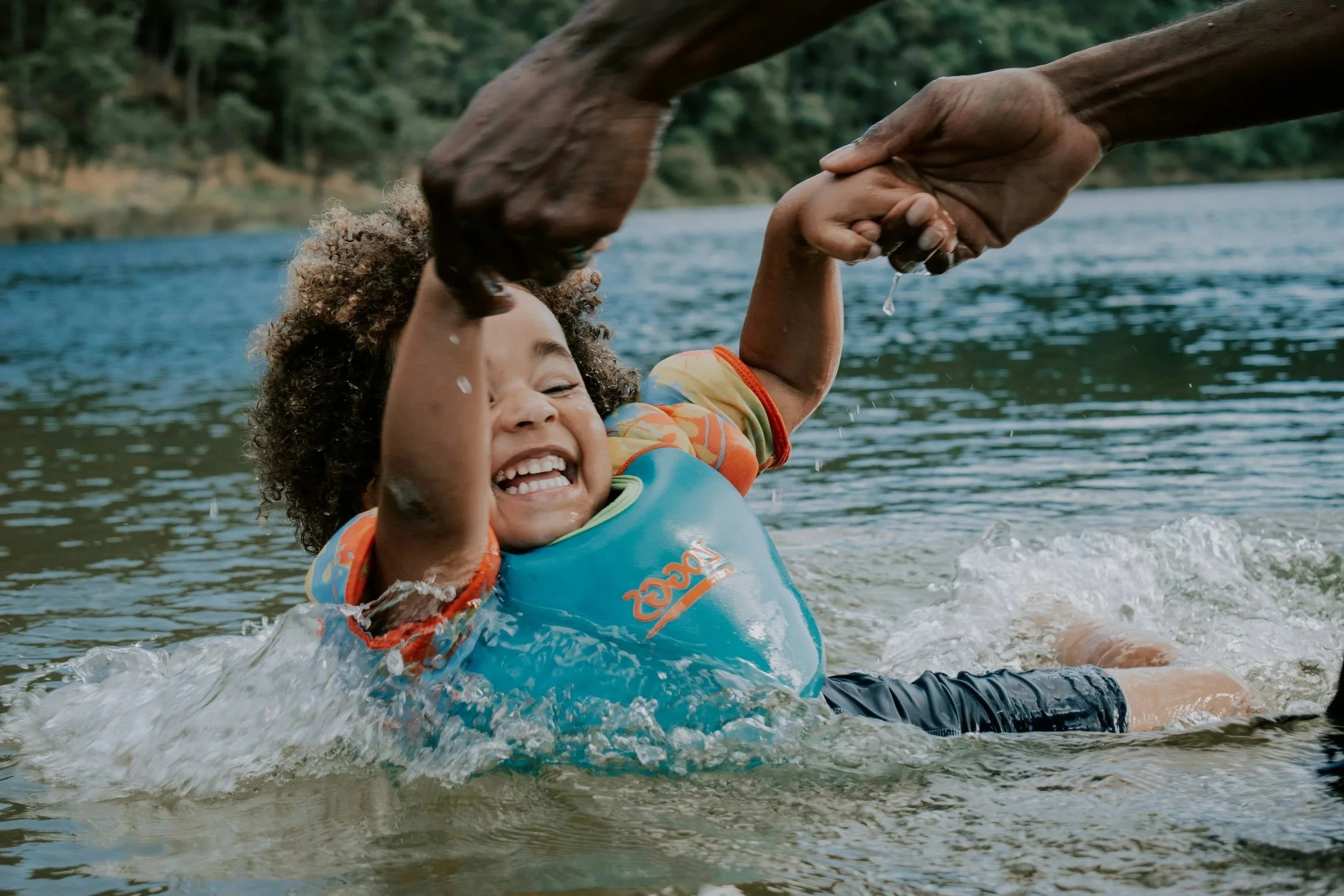 A child with curly hair smiling and enjoying a ride on a canoe or kayak on a river, holding hands with an adult. The child is wearing an orange and blue life jacket and the adult's hand is reaching out to hold the child's hand. In the background, trees line the riverbank.