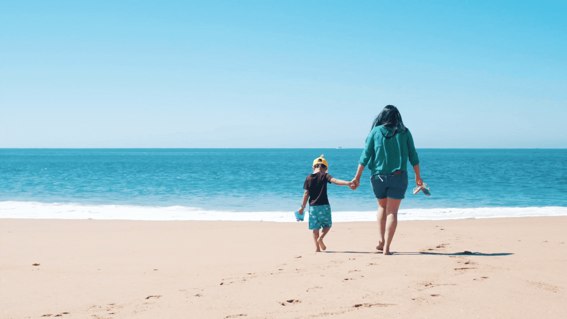 A woman and a child walk hand in hand on a sandy beach towards the ocean, with a clear blue sky above.