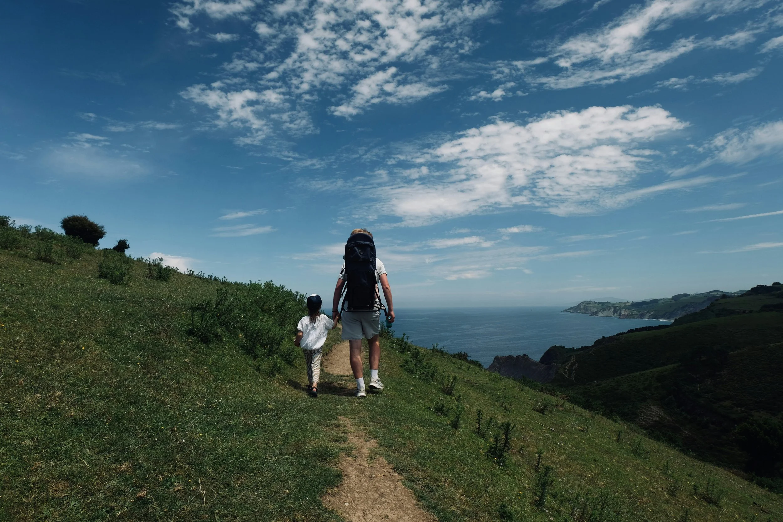 A person and a child walk along a narrow dirt trail on a grassy hillside overlooking the ocean, with blue sky and scattered white clouds above.