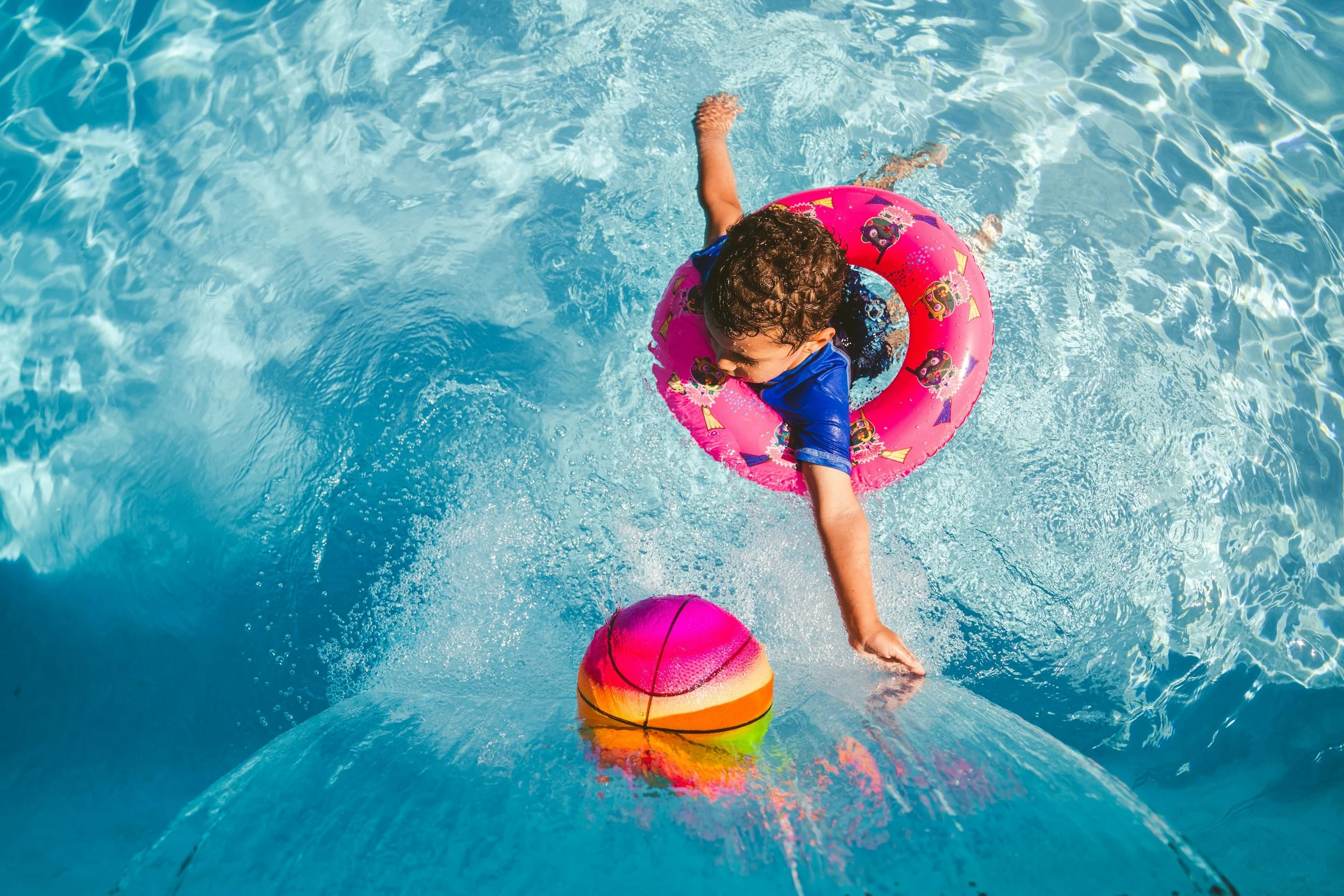 Child swimming in a pool with a pink float ring and colorful ball.