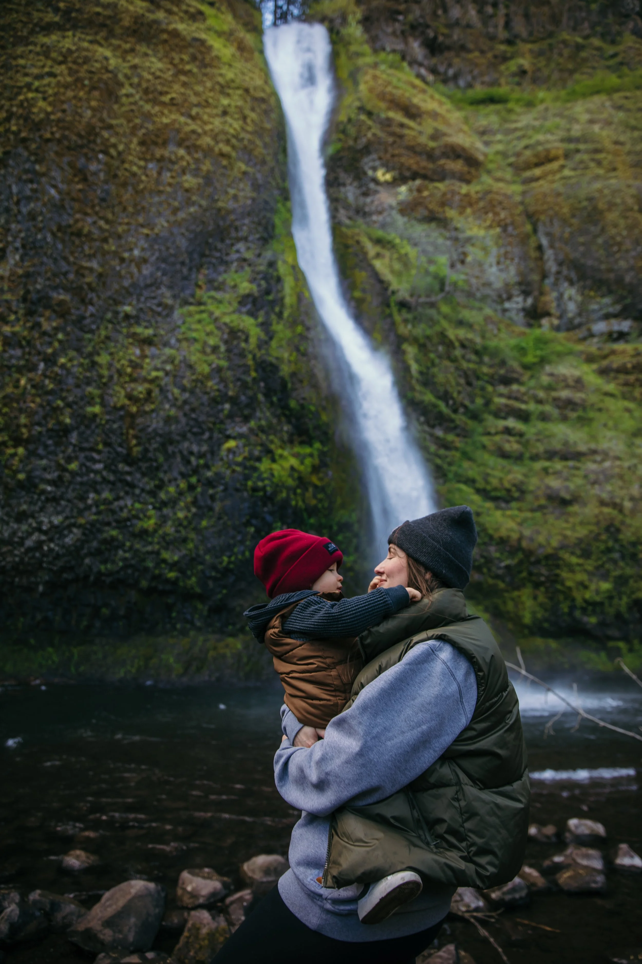 A person carrying a child in front of a waterfall in a lush, green, rocky landscape.