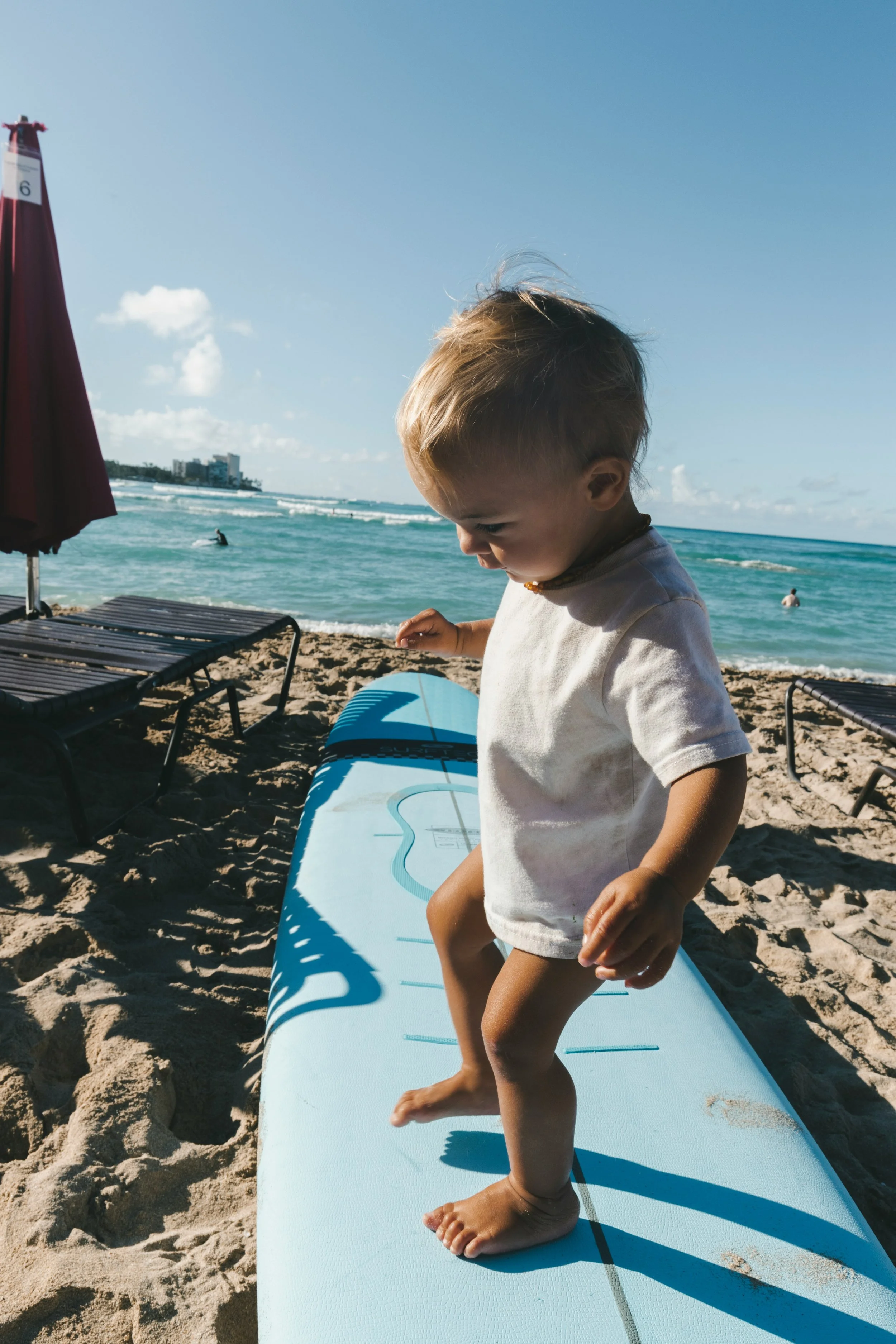 Young child standing on a bright blue surfboard on sandy beach with ocean and blue sky in background.