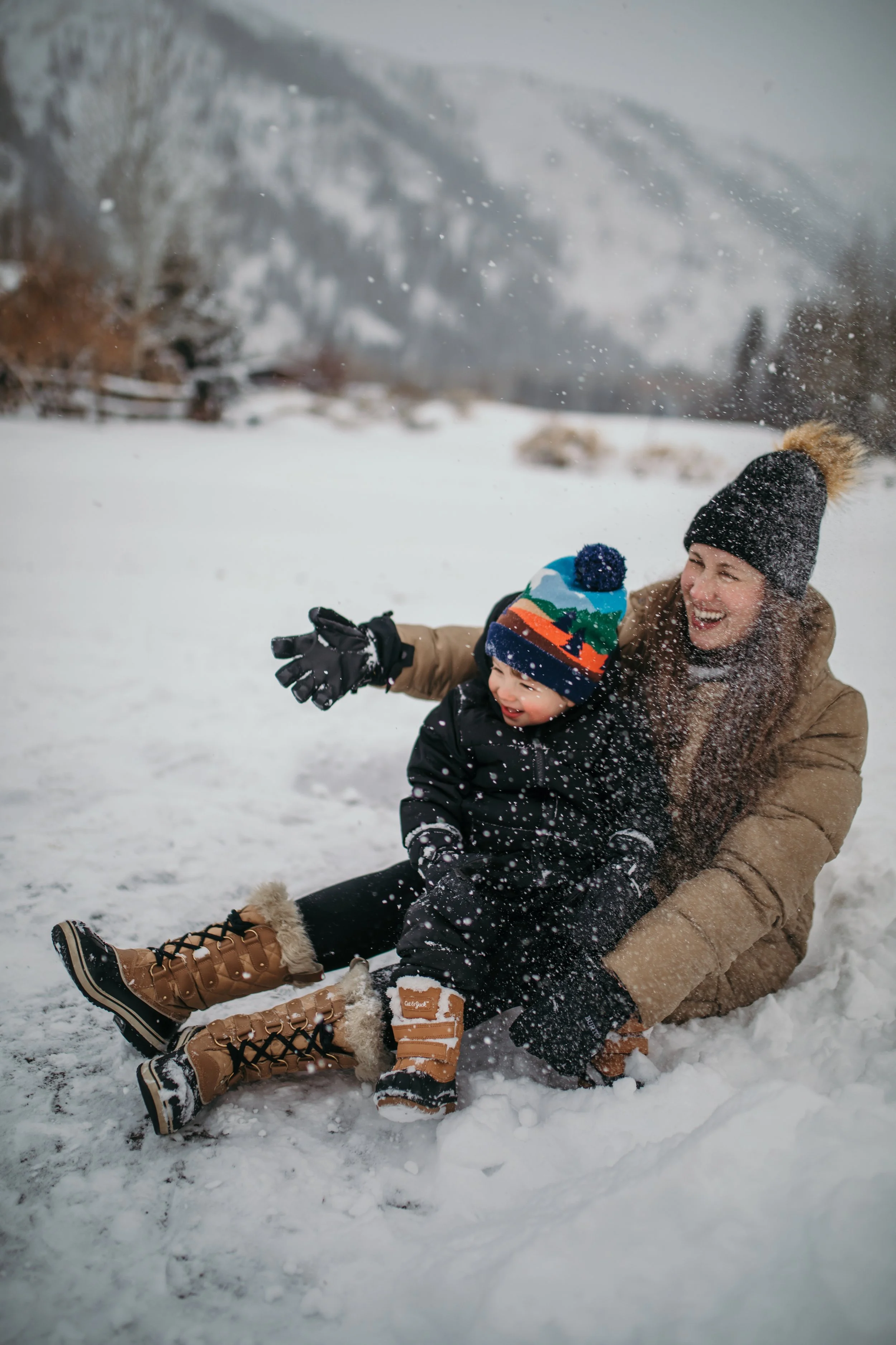 A woman and a young child playing and sitting in the snow, both dressed warmly with winter boots, hats, and gloves, smiling and enjoying a snowy outdoor scene.