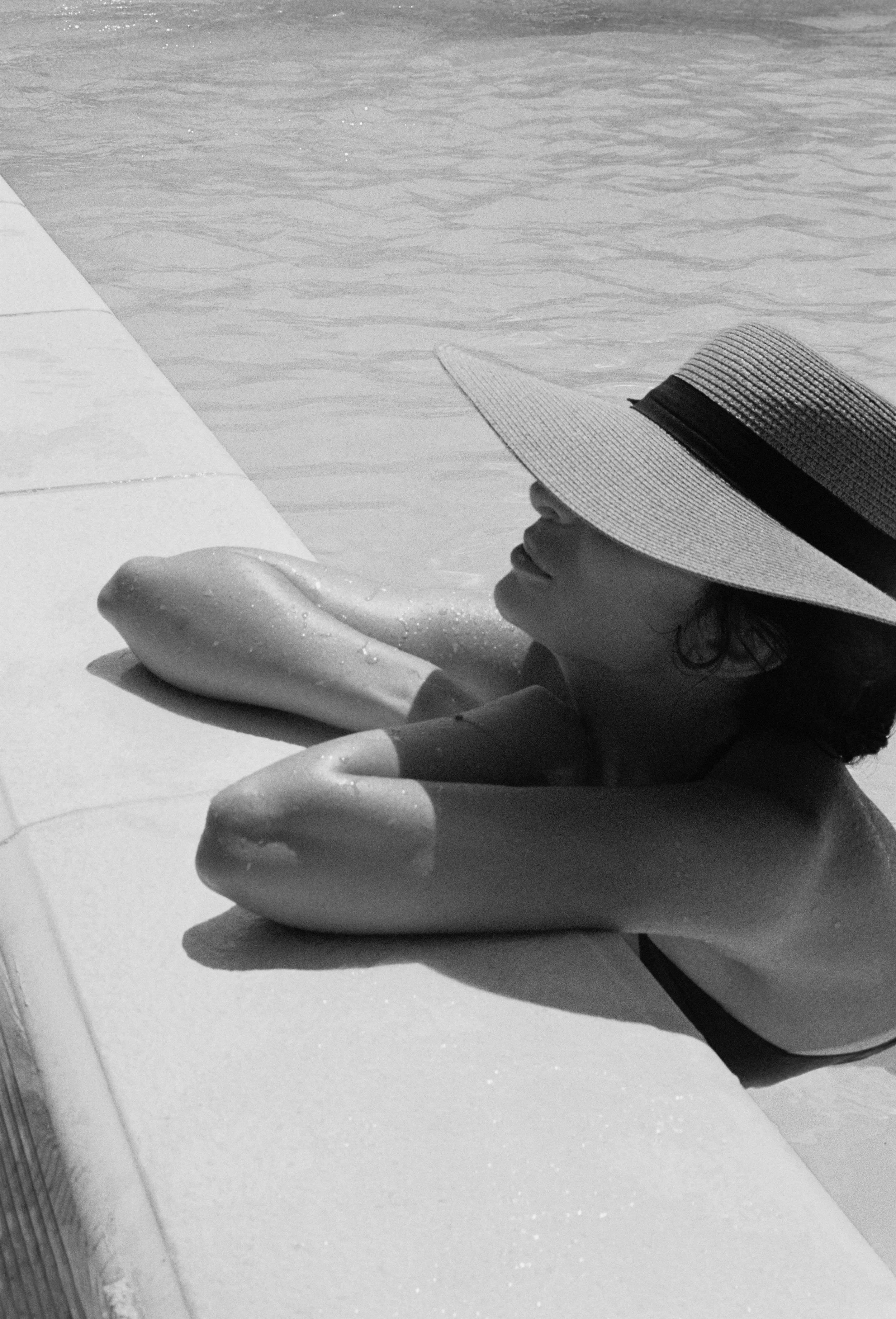 A woman wearing a broad-brimmed hat reclining by a pool, resting her arm on the poolside, with water in the background.