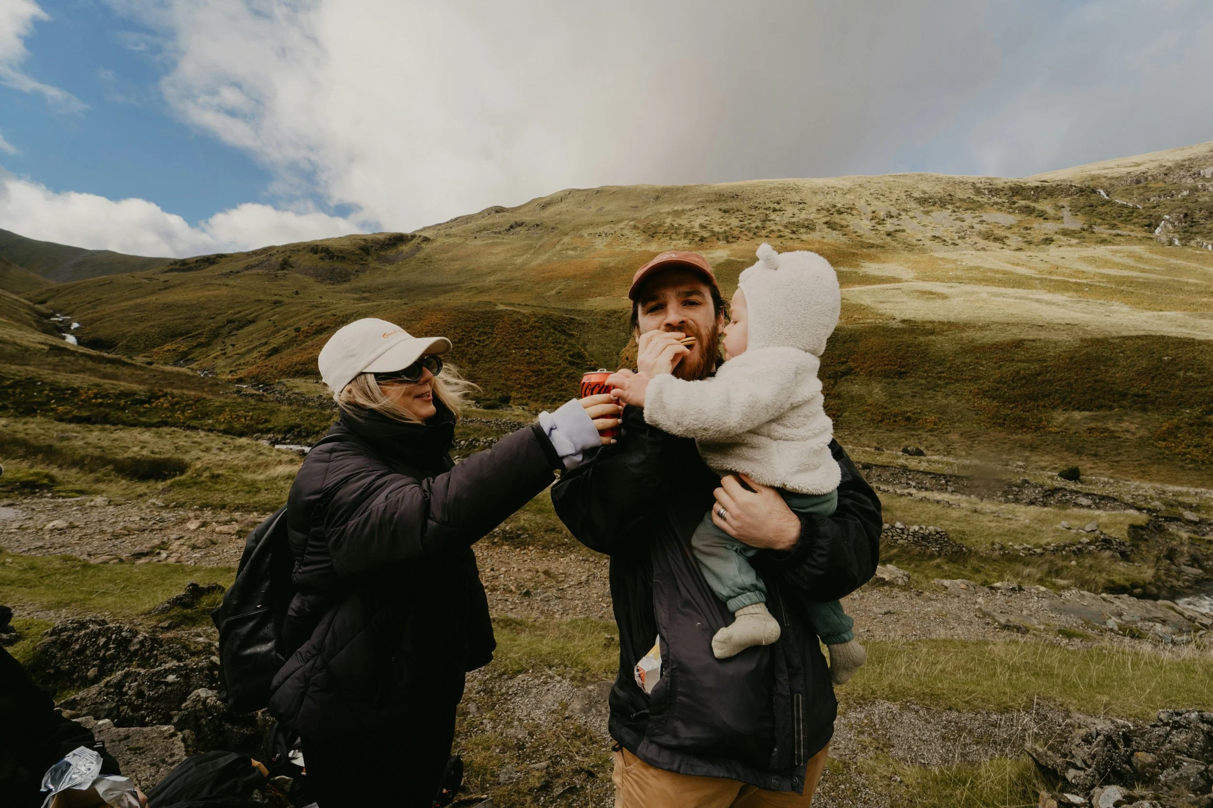 A family of three, consisting of a woman, a man, and a young child, enjoying a moment outdoors in a mountainous landscape. The woman and man are holding the child, who is dressed in a white hoodie, while the man is eating something. The scene features green rolling hills, rocks, and a partly cloudy sky.