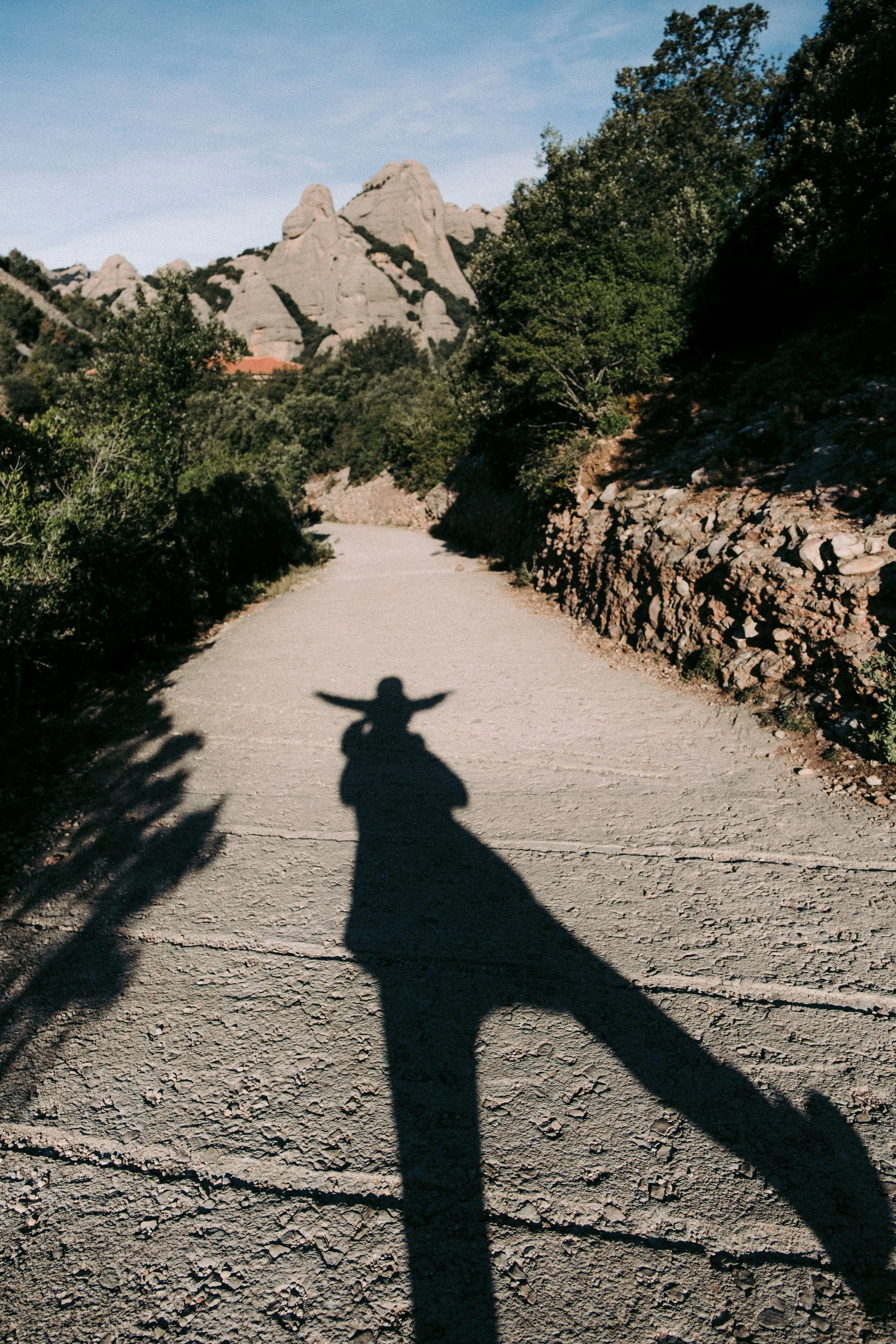 Shadow of a person with arms outstretched on a dirt path, with rocky hills and trees in the background under a clear sky.