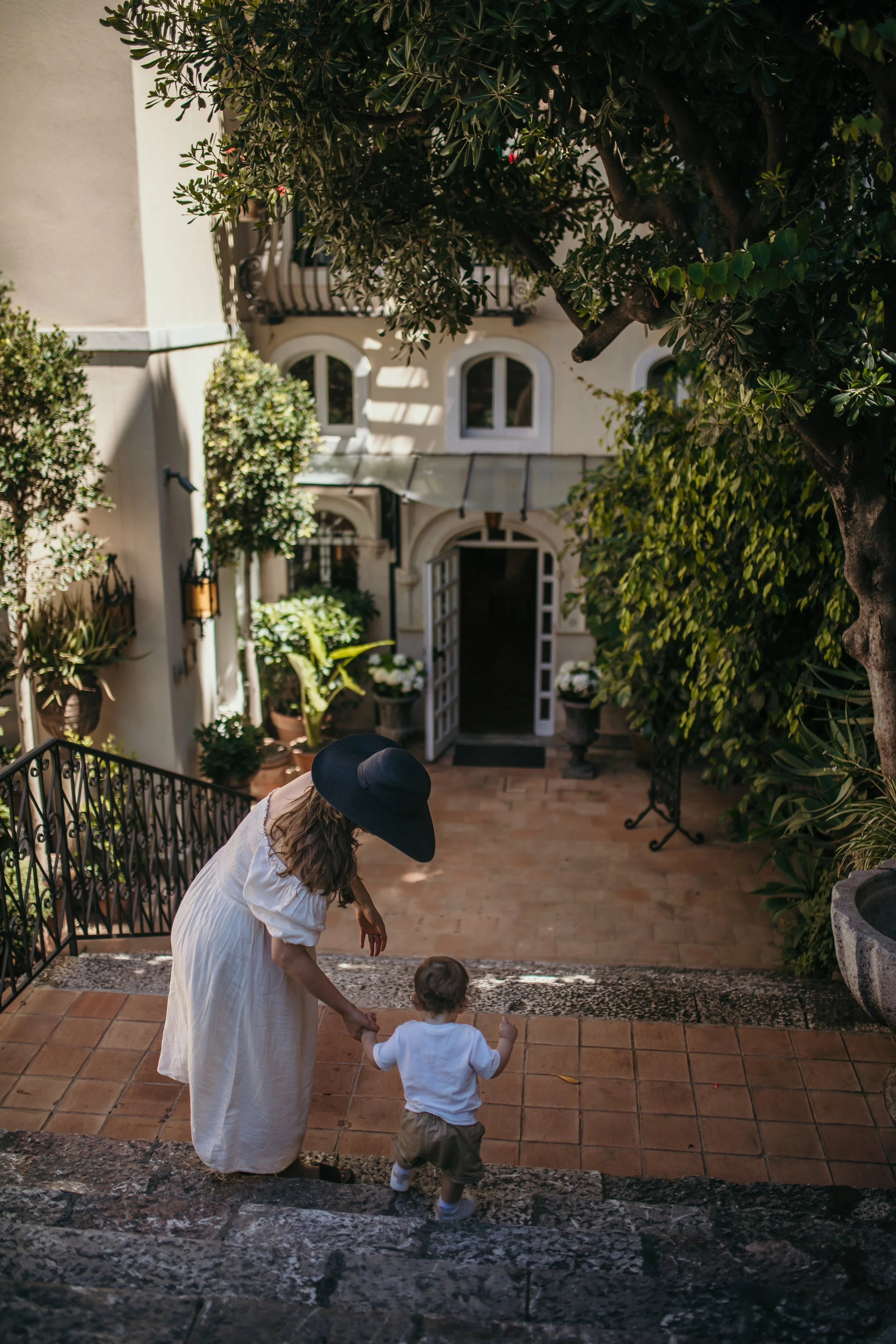A woman and a young boy are descending outdoor stone stairs holding hands, surrounded by lush greenery, in front of a house with arched windows and an open door.