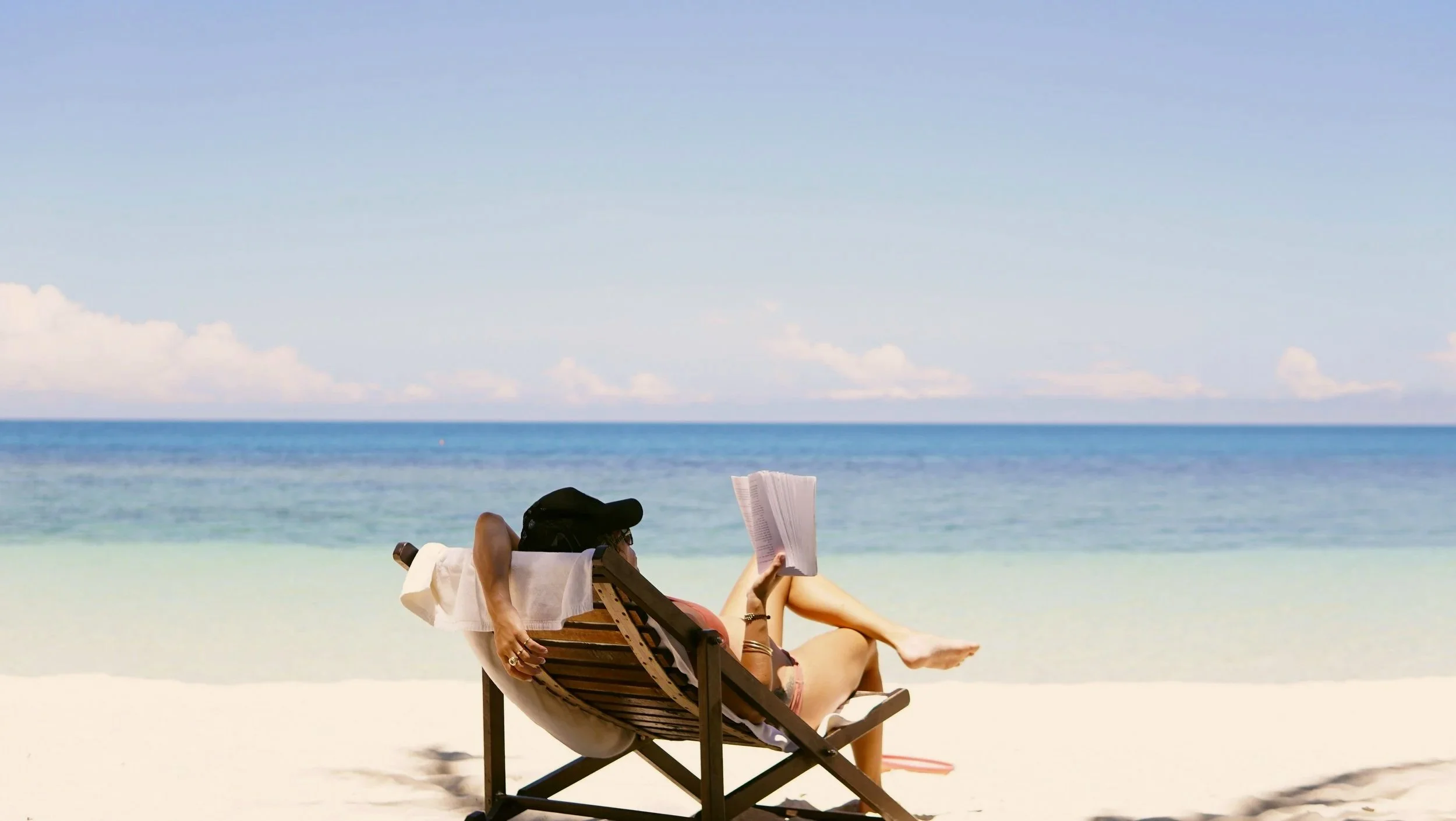 Person relaxing on a lounge chair at the beach, reading a book with the ocean in the background.