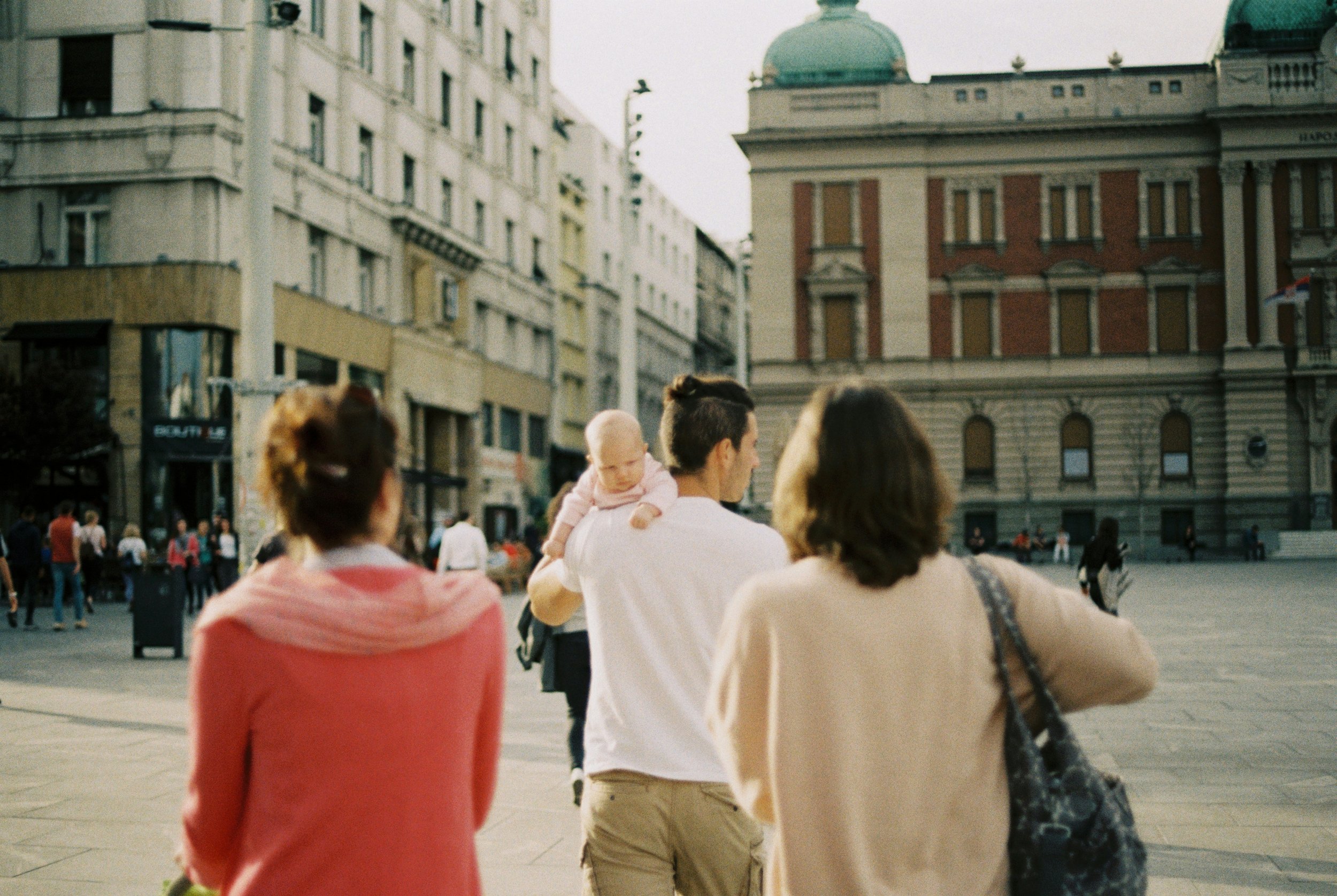 A group of people walking in a busy city square with historic buildings in the background. One person is carrying a baby.