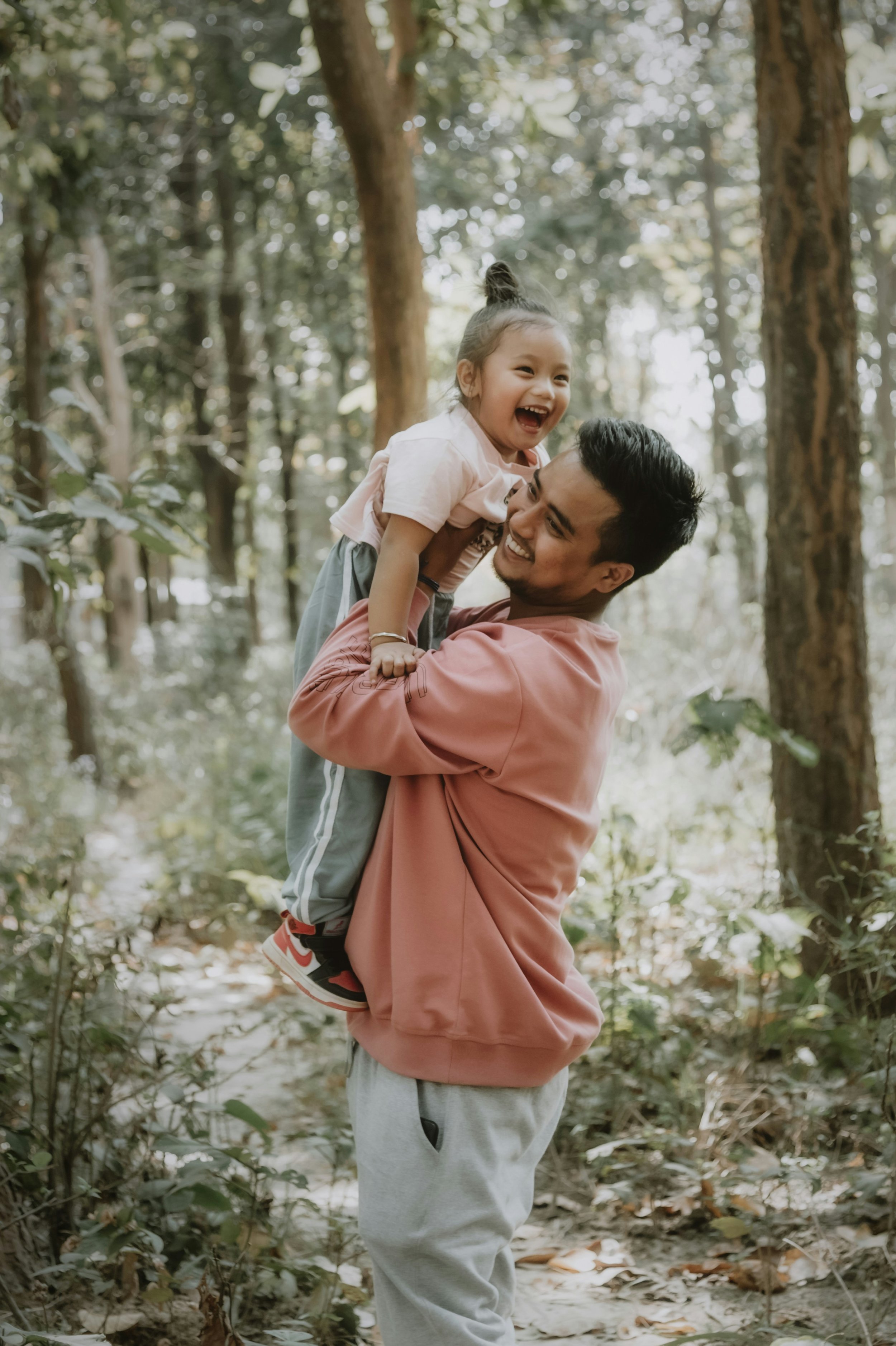 A man lifting a young girl in a forest, both are smiling and enjoying time together among trees and greenery.
