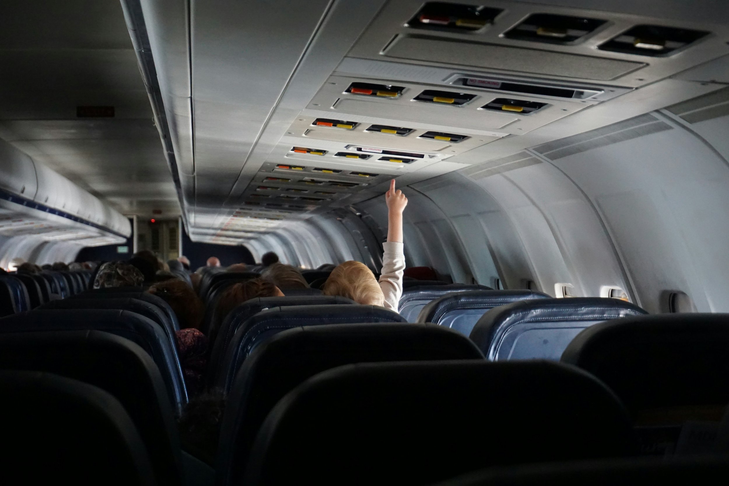 Inside an airplane cabin with passengers seated, one person raising their hand near overhead compartments.