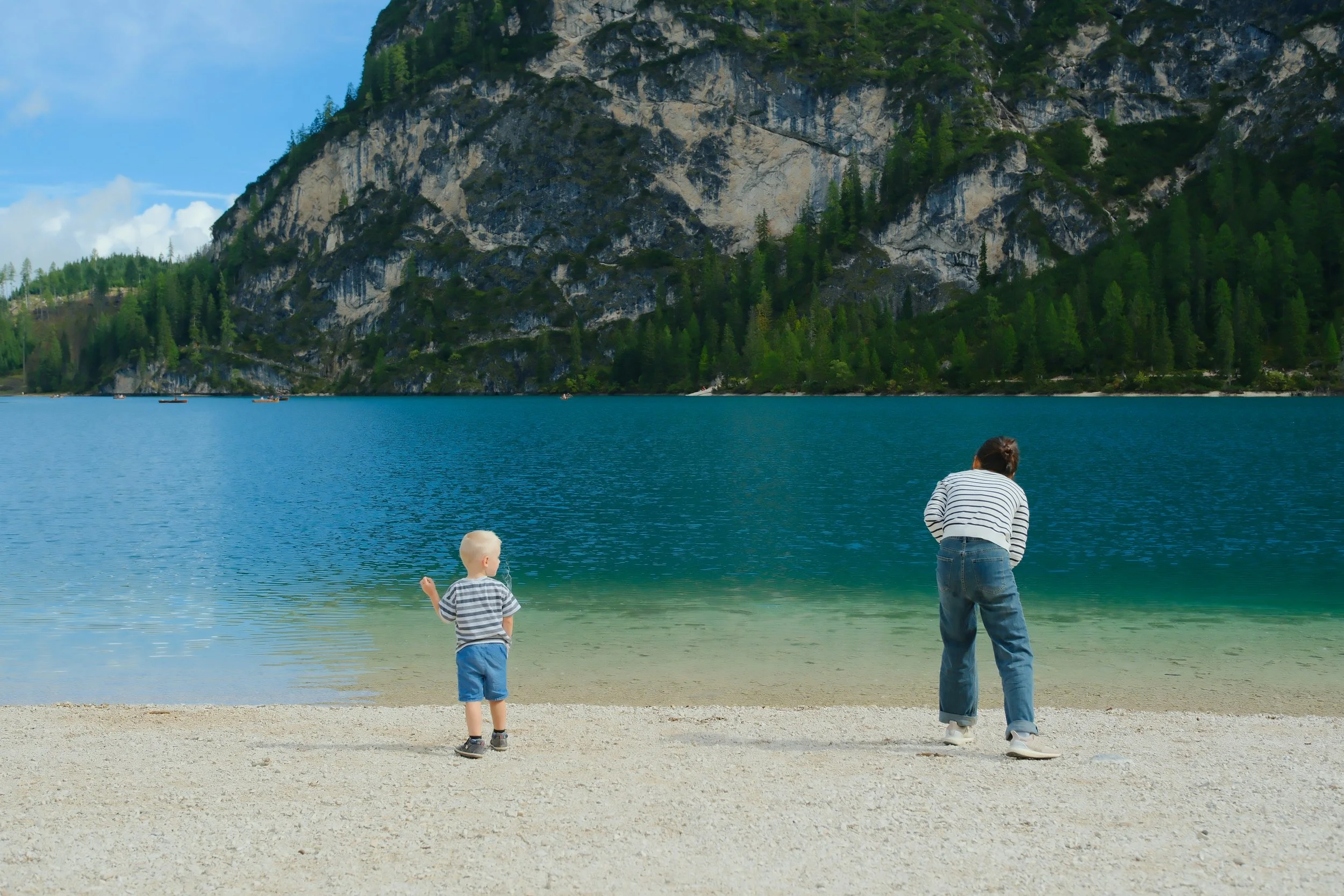 A woman and two young boys on a sandy beach near a lake with mountains and trees in the background. The woman and boy are facing the water while the other boy is looking towards the water.