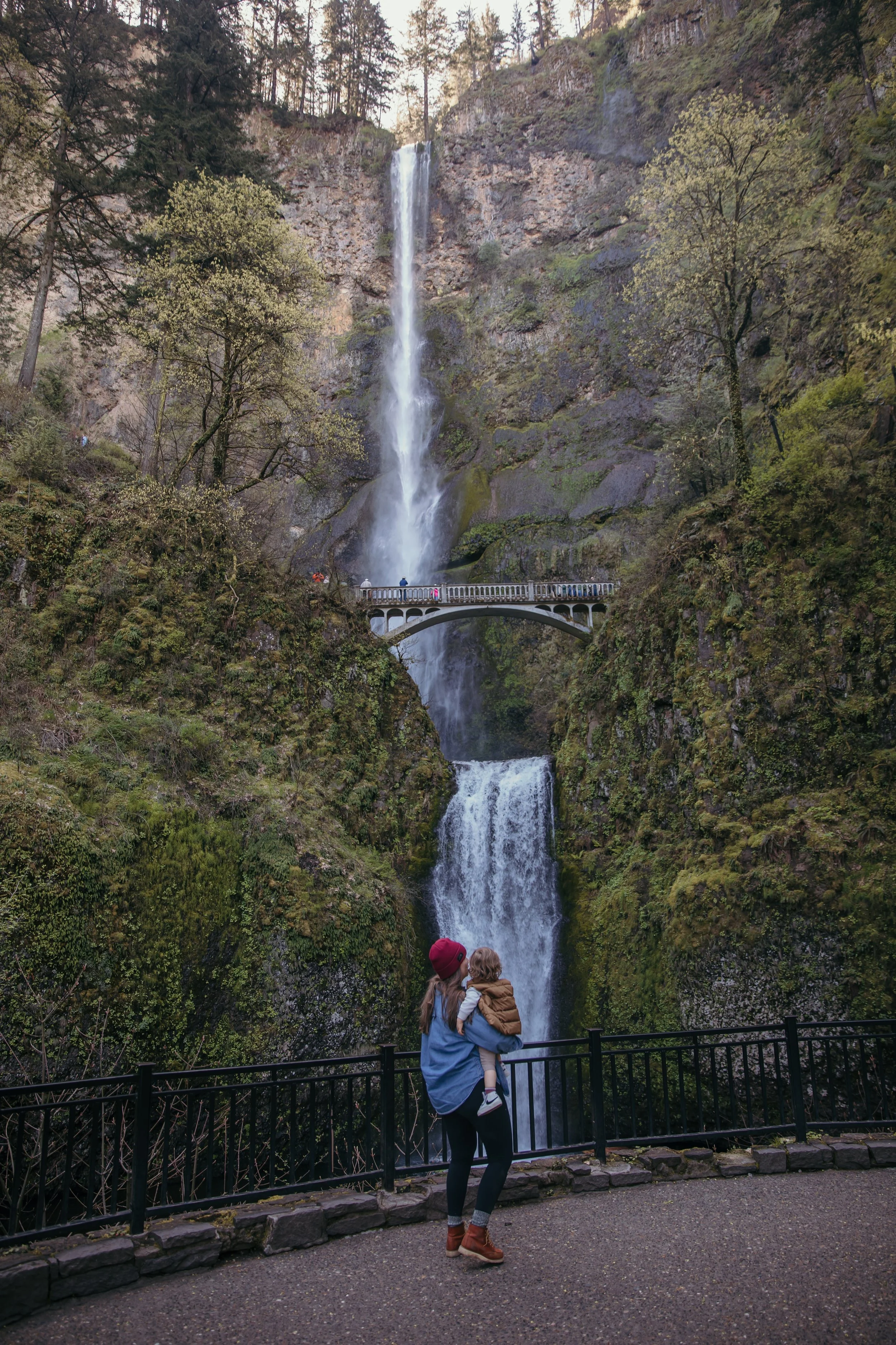 A woman holding a child while looking at Multnomah Falls, a tall waterfall in a lush forest with a bridge across the waterfall.