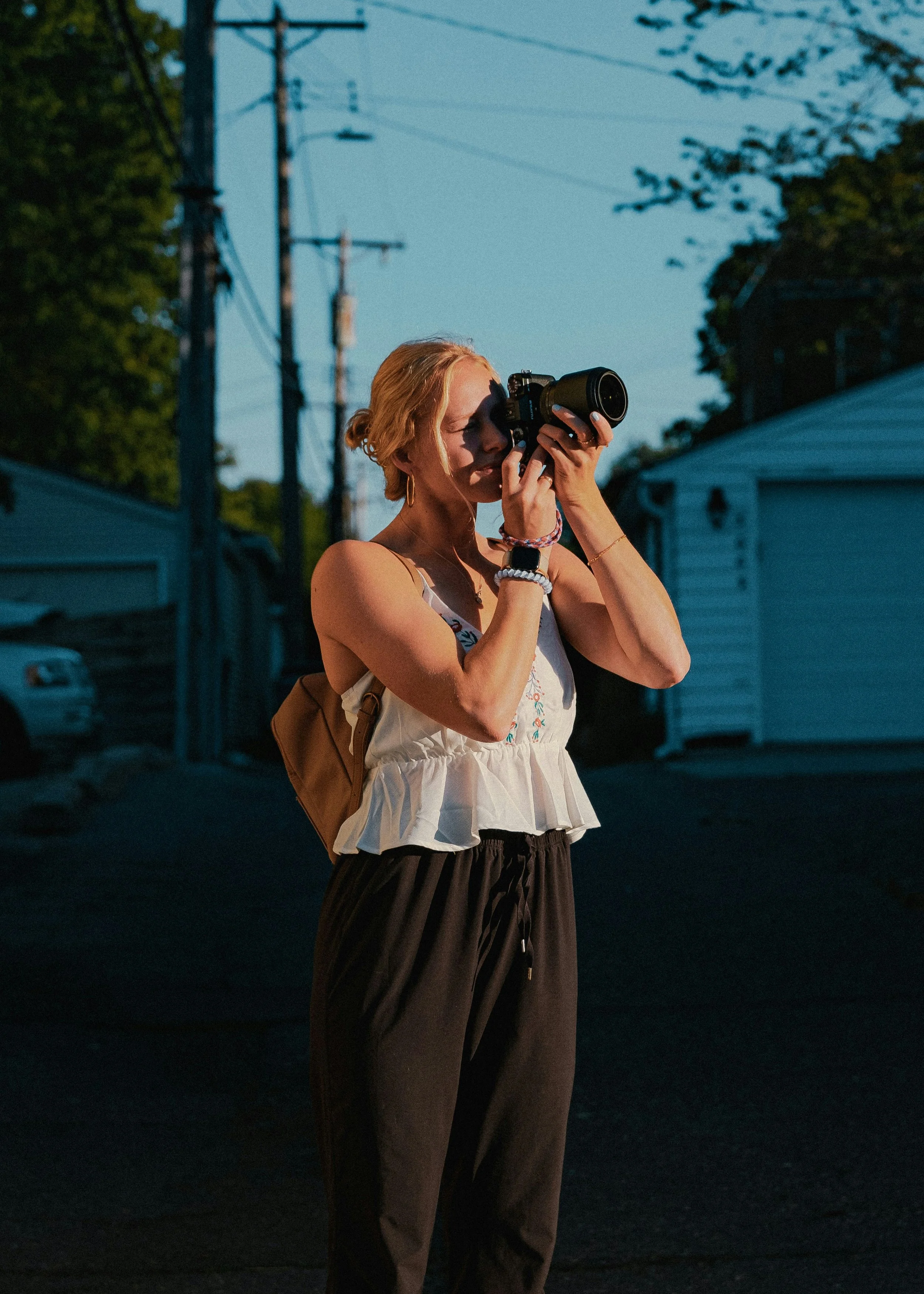 A woman with blonde hair taking a photo with a camera outdoors during the evening, wearing a white top and brown pants, with trees and power poles in the background.