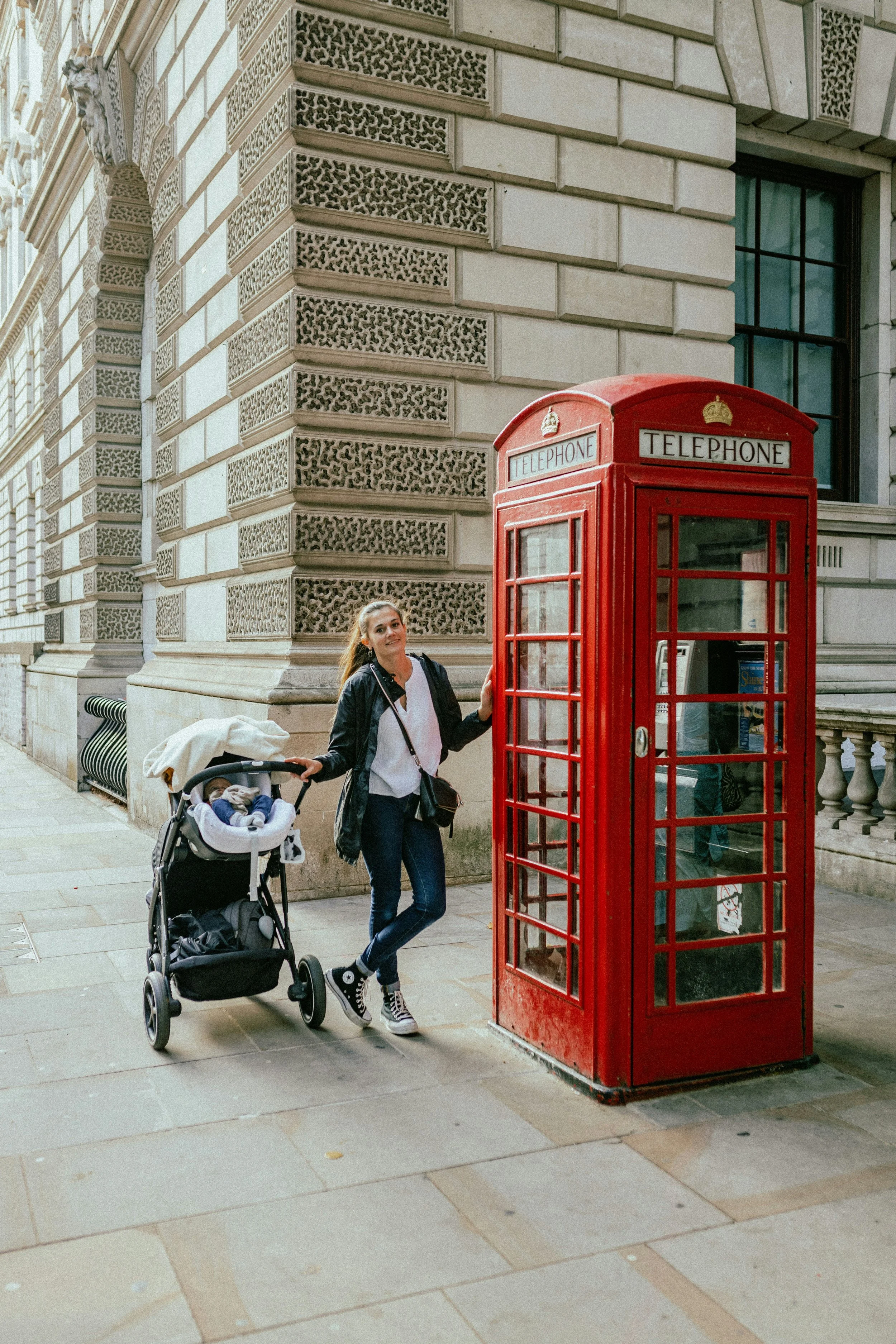 A woman with long hair in a ponytail, wearing a black jacket, white shirt, and sneakers, stands next to a red British telephone booth. She is holding a stroller with a sleeping baby inside. The scene is outdoors in front of a historical building with detailed stonework and a large window.