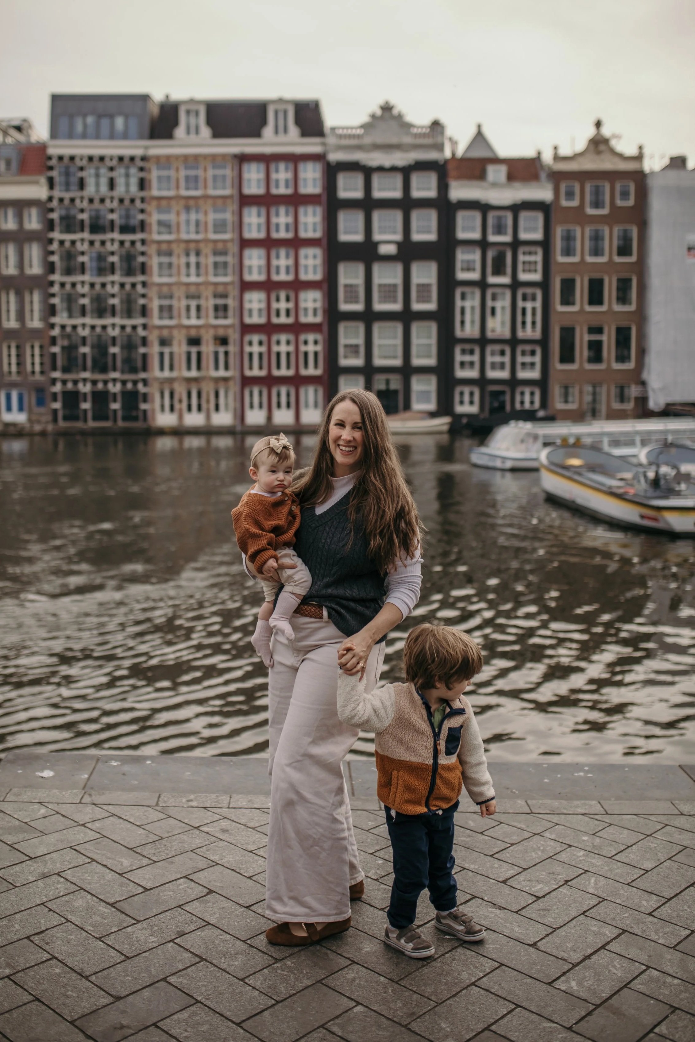 A woman holding a young girl by the hand, standing near a body of water, with colorful historic buildings and boats in the background.
