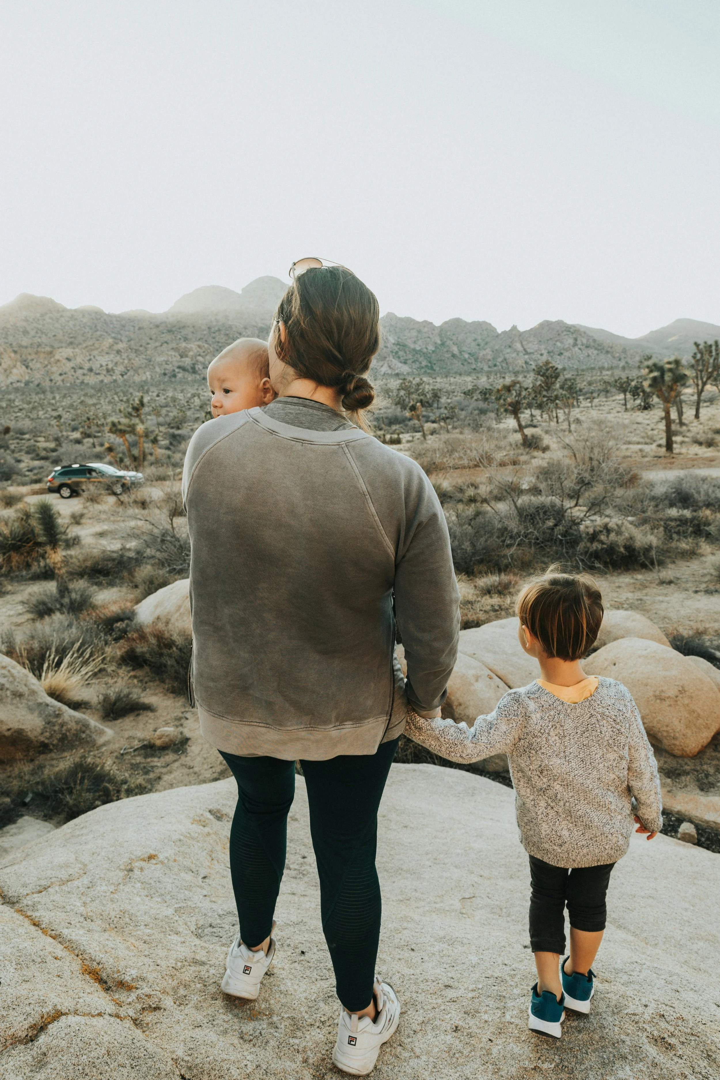 A woman and two children holding hands and walking through a desert landscape with mountains and Joshua trees in the background. The woman is holding a baby and the older child is walking beside her.