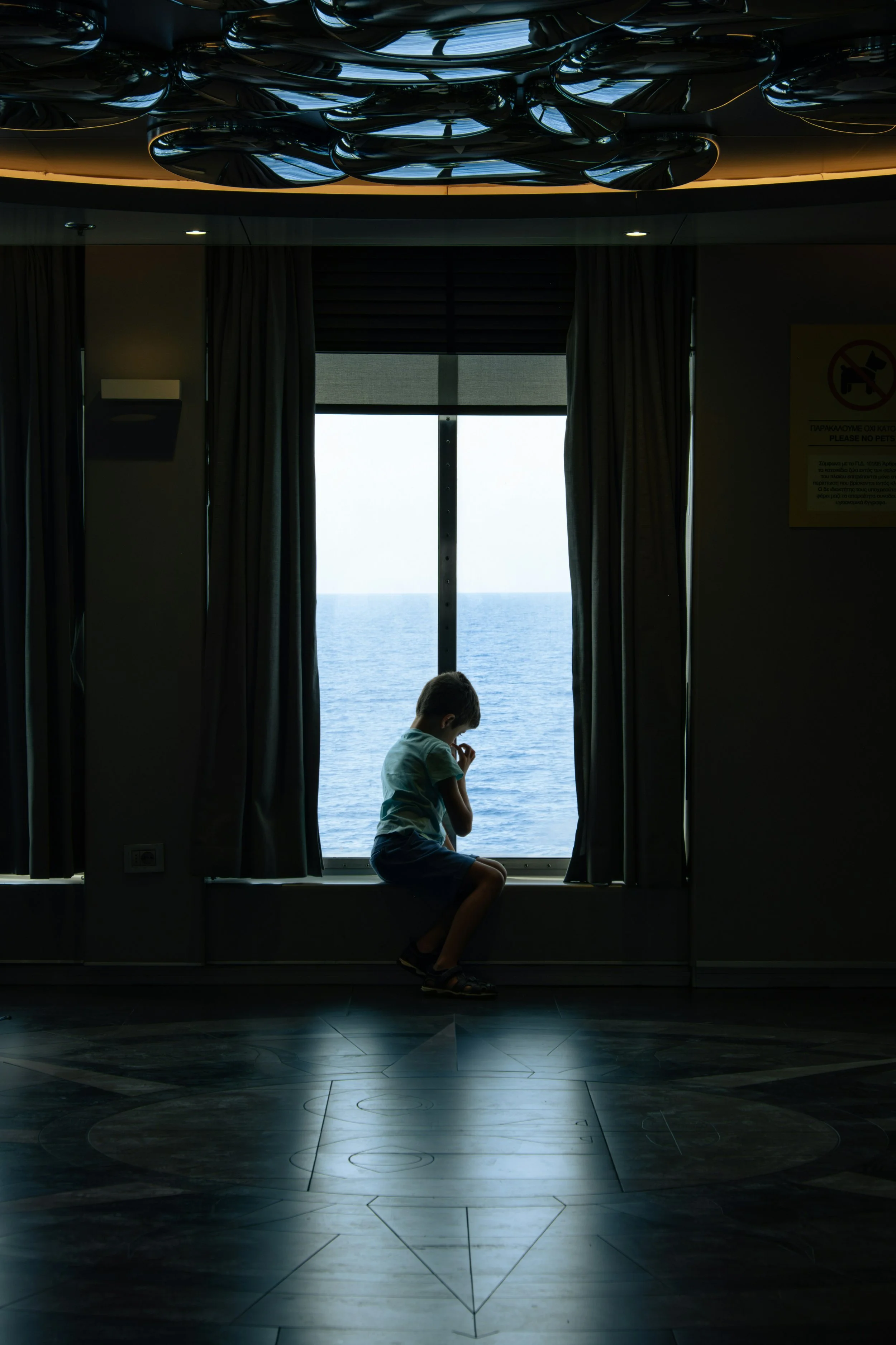 A young boy is sitting on the windowsill of a large window with dark curtains pulled to the sides, overlooking a body of water. The boy appears to be looking down, with his hand near his mouth. The room is dimly lit with some ceiling lights, and there is a sign on the wall to the right.