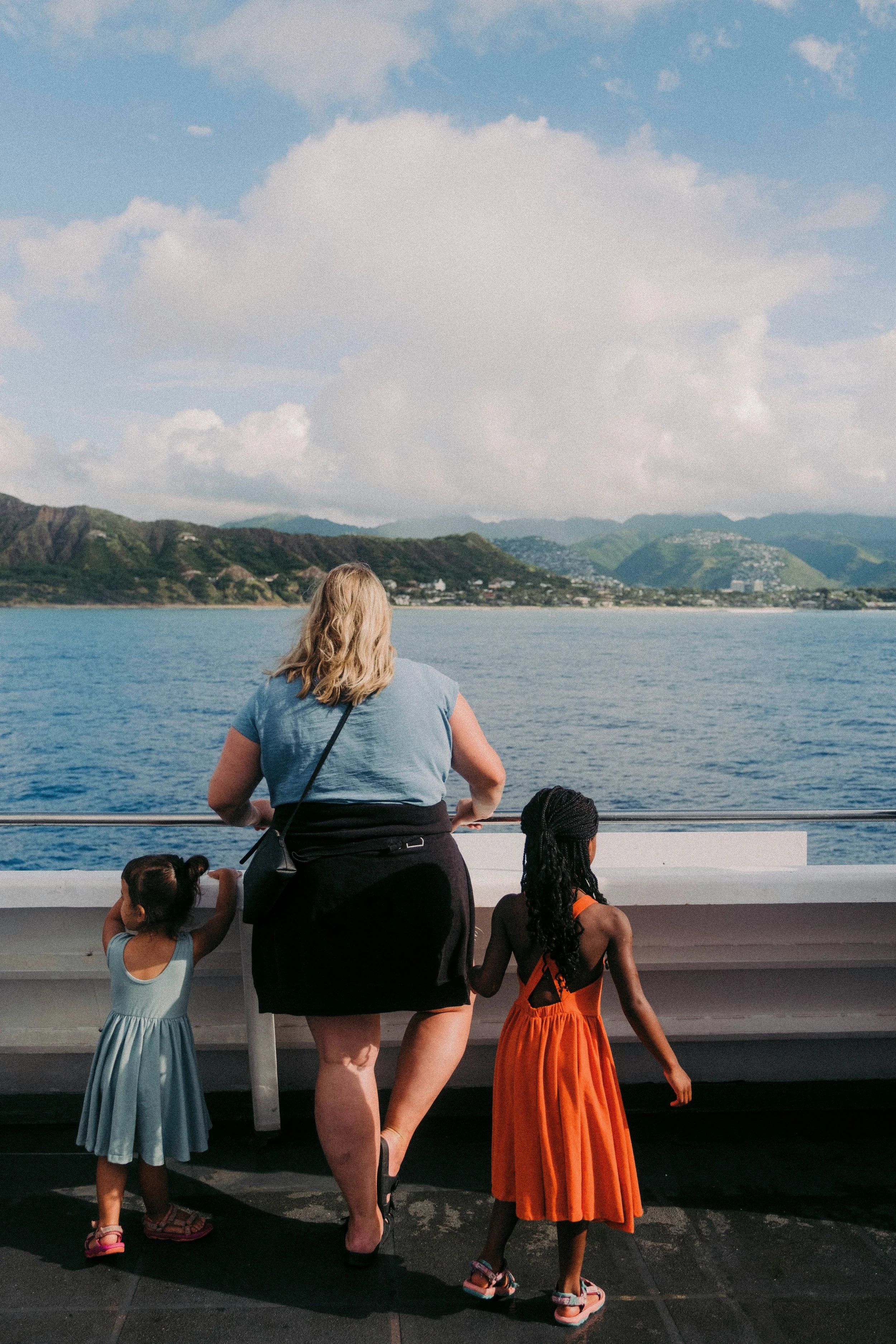 A woman and three children standing on a boat deck, looking at the water and mountains in the distance. The woman is wearing a light blue shirt and black skirt with sandals, and has blonde hair. The children are wearing colorful dresses and sandals, with hairstyles including braids and curly hair. The water is blue, and the sky is partly cloudy.