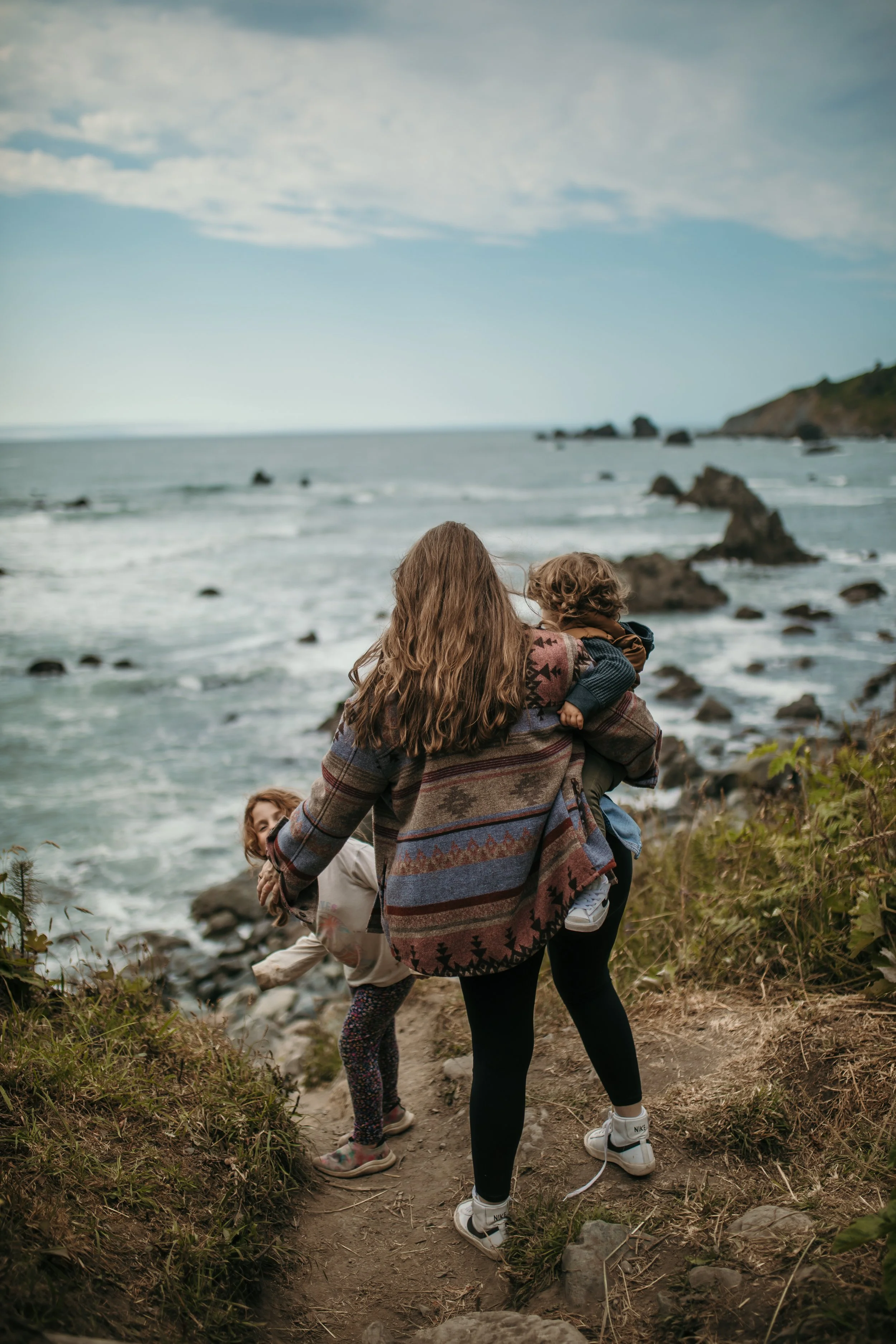 A woman holding a child on her back walking along a rocky and grassy trail by the coast, with another child nearby reaching down to pick up something from the ground, and the ocean with large rocks and a cloudy sky in the background.