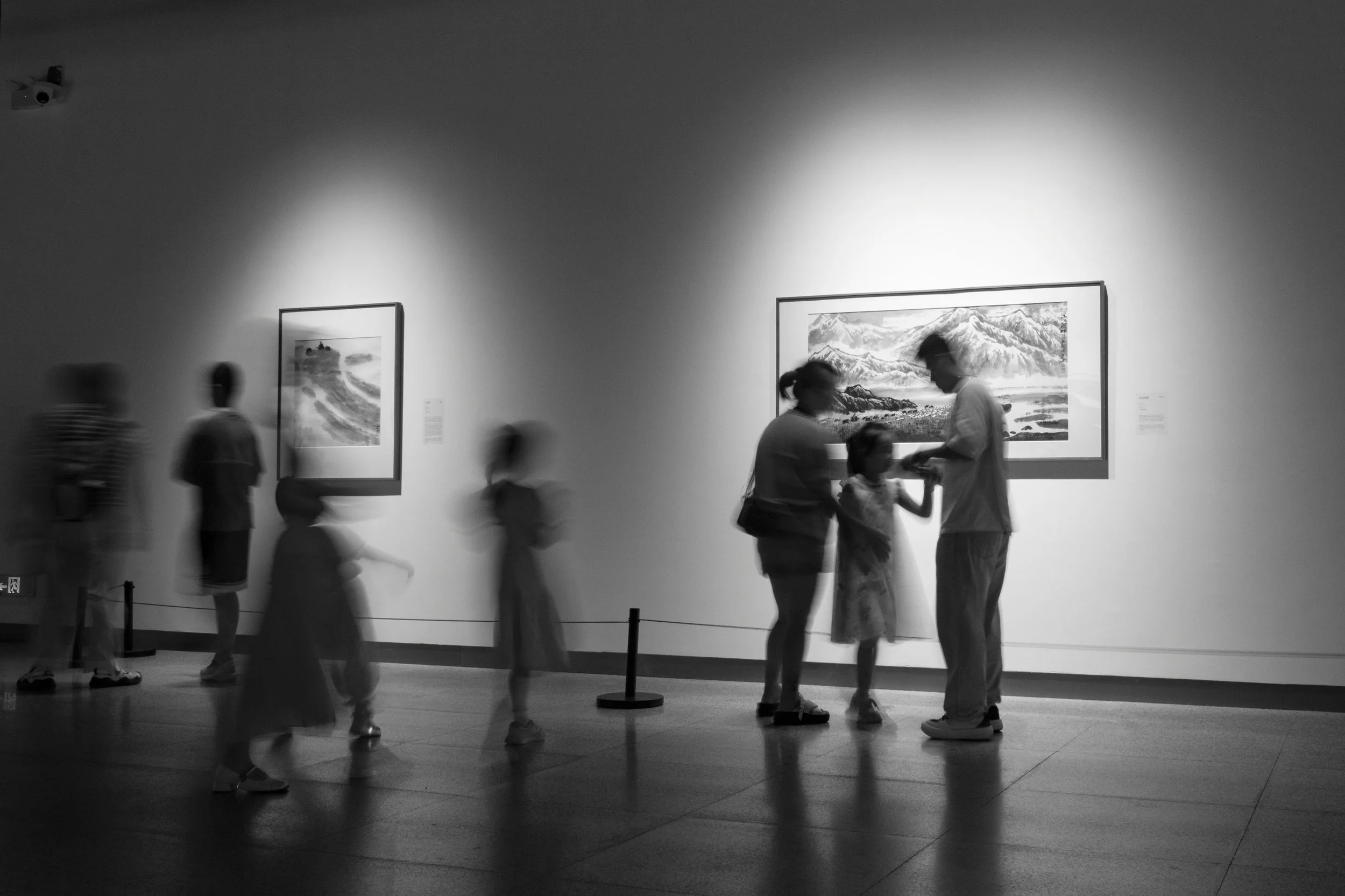 People viewing artwork in a museum gallery, including framed pictures on the wall and individuals engaging with the exhibits.