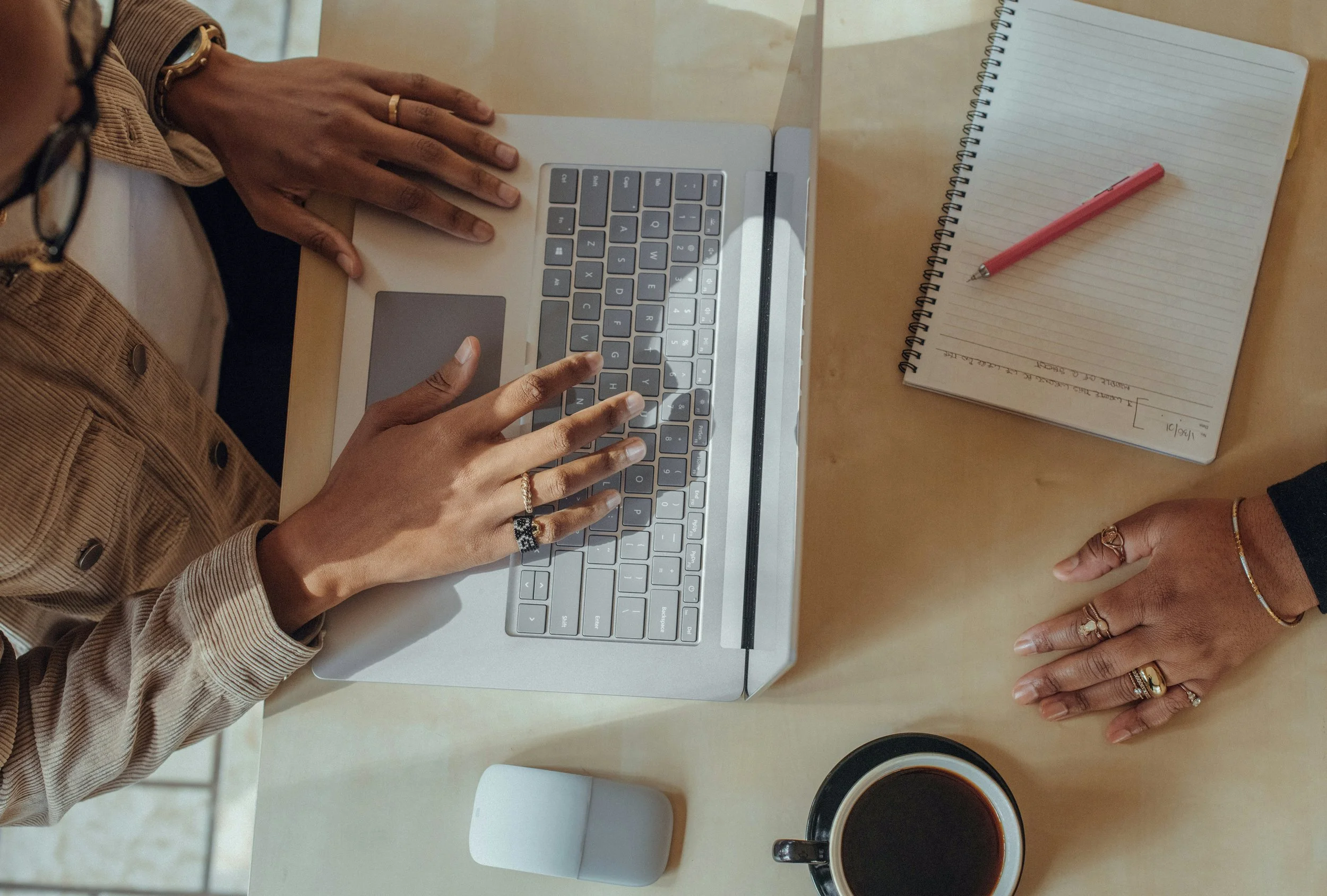 A person with medium-dark skin typing on a silver laptop with a gray keyboard. The person is wearing rings, a beige jacket, and a watch. On the wooden table, there is an open spiral notebook with lined pages and a pink pen, and a black coffee mug filled with coffee.