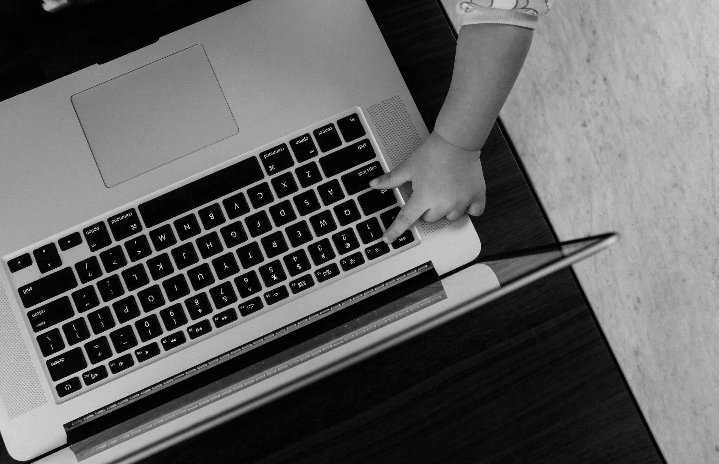 Black and white photo of a child pointing at a laptop keyboard with their index finger, lying on a wooden table with a textured surface, and a paper to the left.
