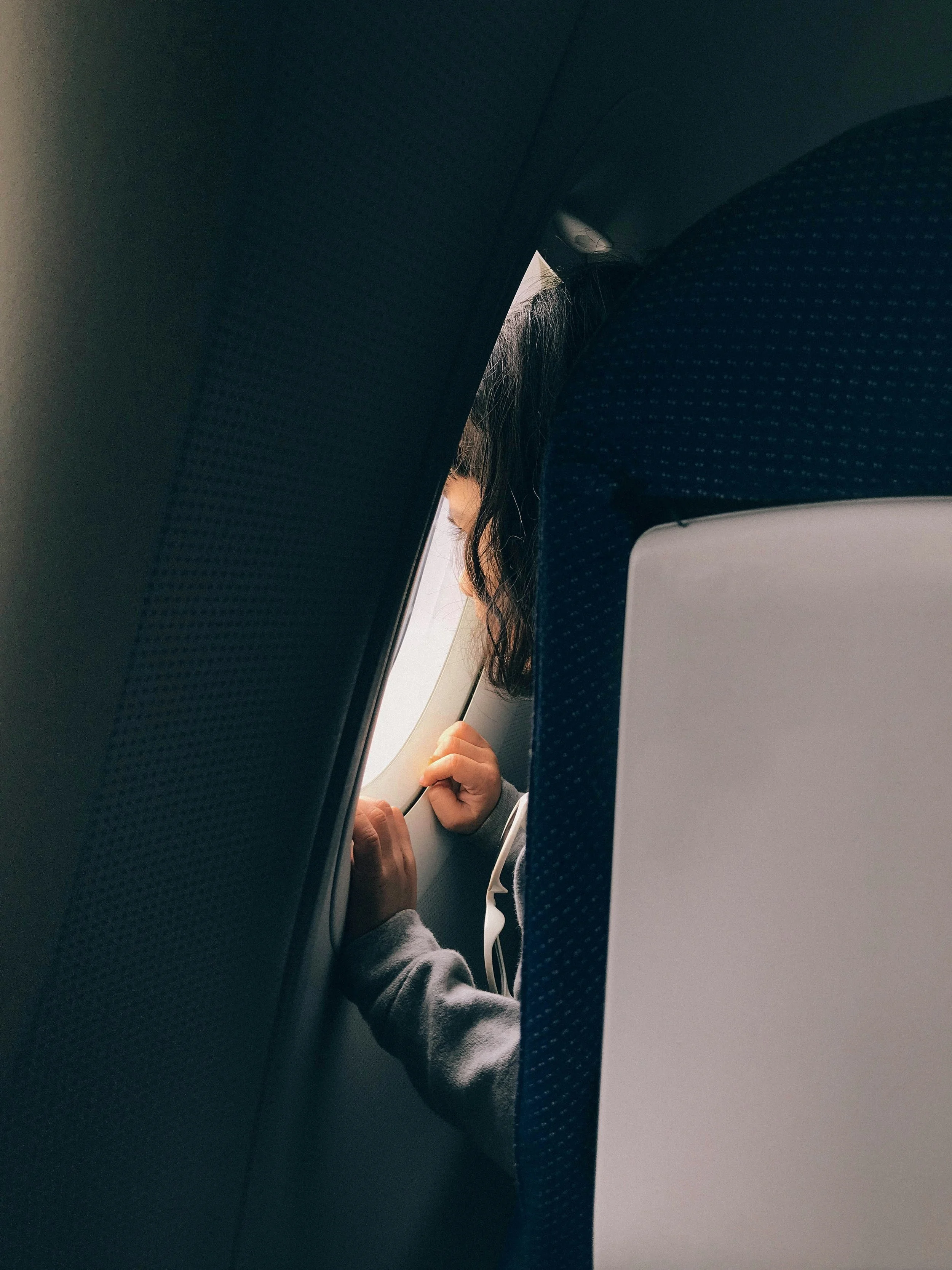 Person with dark hair and light skin sitting on an airplane, looking out the window, hand resting on the armrest, wearing a gray hoodie.